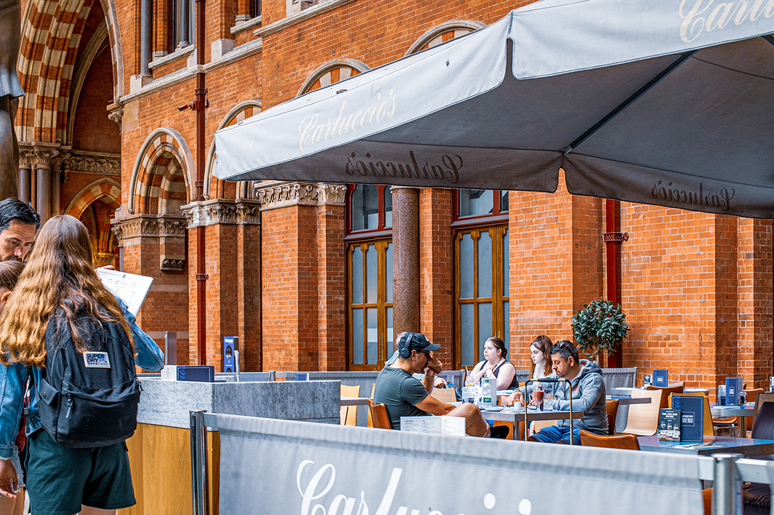 The image depicts an outdoor seating area of a café or restaurant named 'Carluccio's'. The setting is in front of a brick building with large arched windows and ornate columns. Several people are seated at tables under large umbrellas, enjoying their meals and drinks. Two individuals in the foreground appear to be looking at a menu or a map. The atmosphere seems relaxed and casual, typical of a pleasant day at an outdoor dining spot.