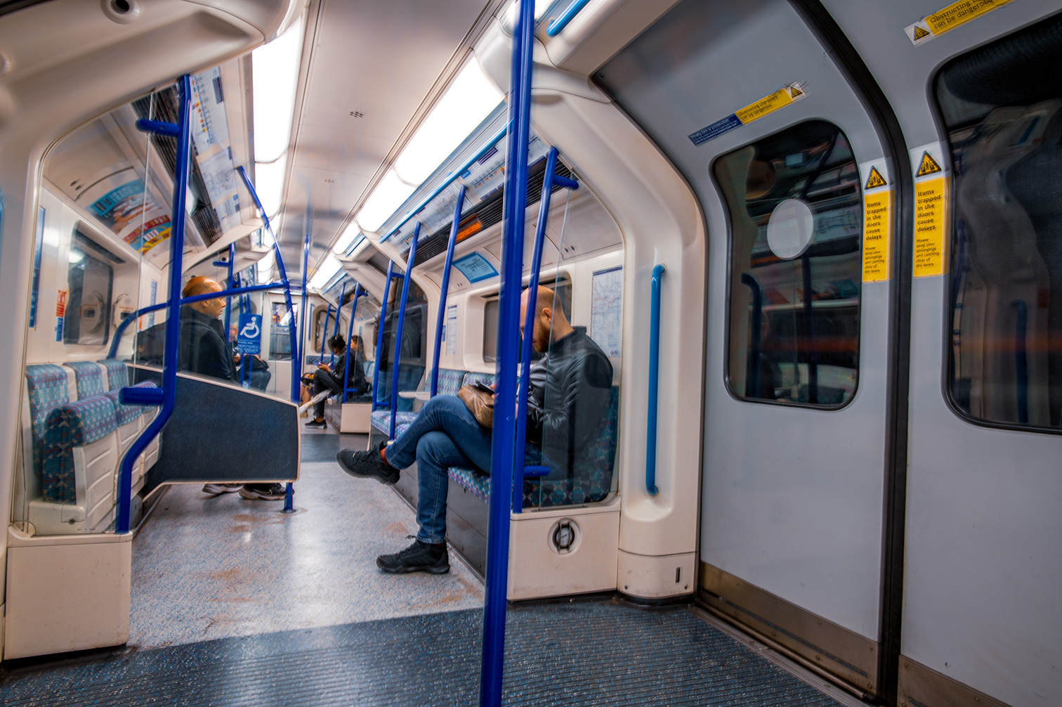 The image depicts the interior of a modern subway train. The train is relatively empty, with only a few passengers visible. The passengers are seated and appear to be engaged in various activities such as looking at their phones or resting. The train has blue handrails and seats with a patterned design. Safety notices are prominently displayed on the walls near the doors.