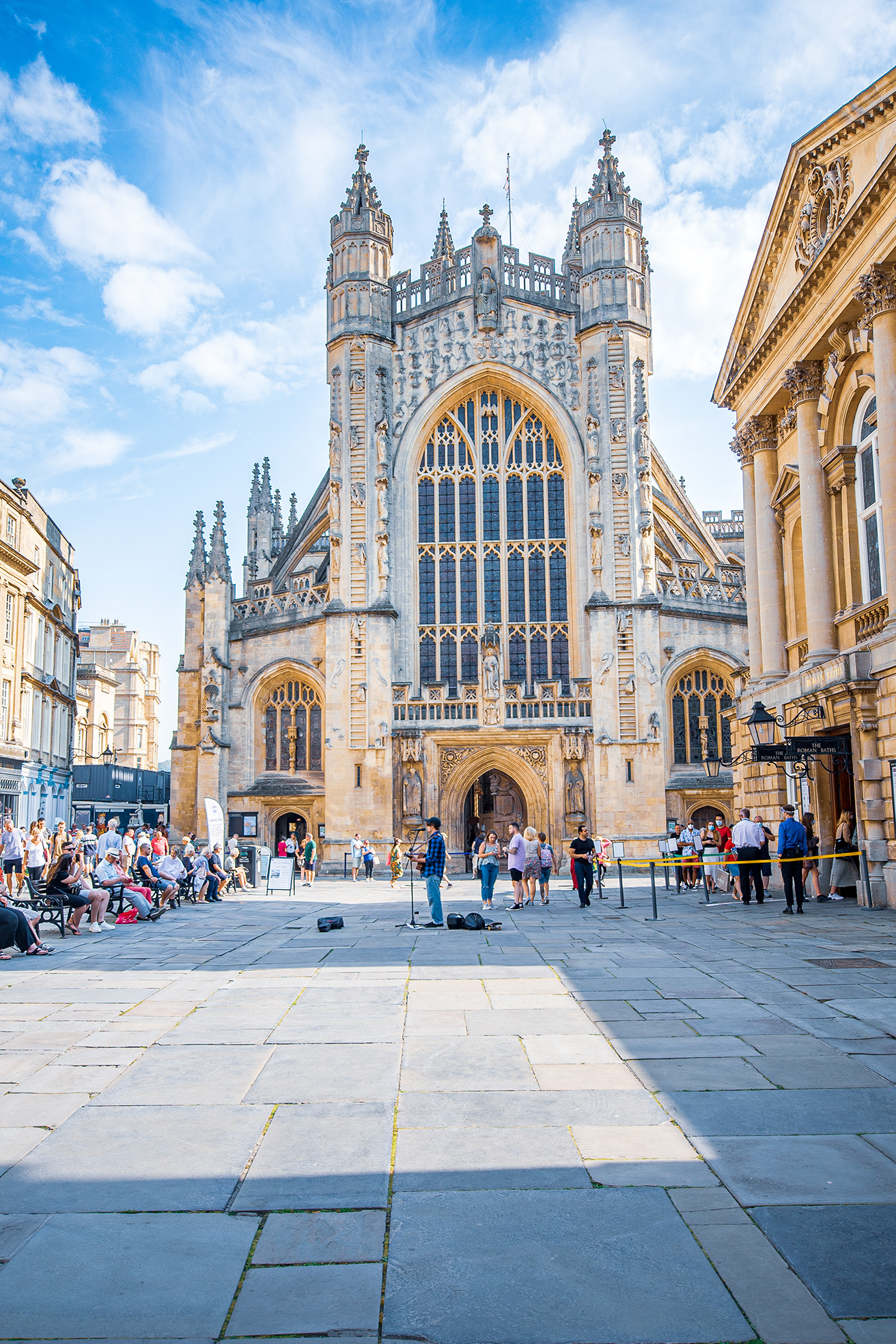 The image depicts a bustling scene in front of a large, ornate Gothic-style building, likely a cathedral or church, with intricate architectural details and large stained-glass windows. The sky is clear with a few clouds. People are gathered in the square, some sitting on benches, others standing or walking around. A street performer is playing a guitar in the foreground. The overall atmosphere is lively and vibrant, with tourists and visitors enjoying the historic and architectural beauty of the location.