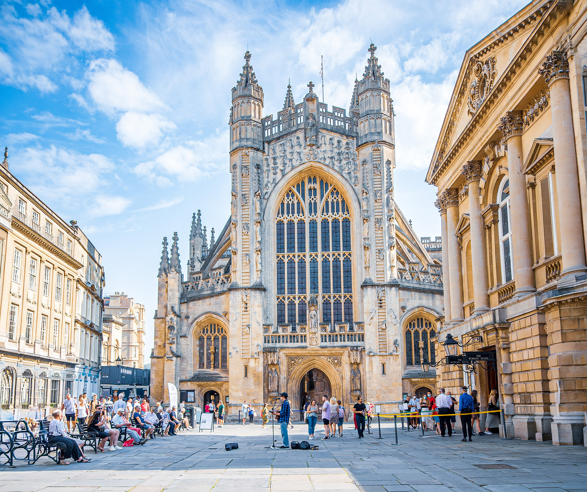The image depicts a bustling scene in front of a grand, historic building, likely a cathedral or church, characterized by its large arched window with intricate tracery and two tall, ornate towers. The architecture is Gothic, with detailed stone carvings and pointed arches. The building is situated in a square with people sitting on benches, walking around, and engaging in various activities. The sky is clear with a few clouds, suggesting a pleasant day. The surrounding buildings have a similar historic architectural style, contributing to the overall aesthetic of the scene.