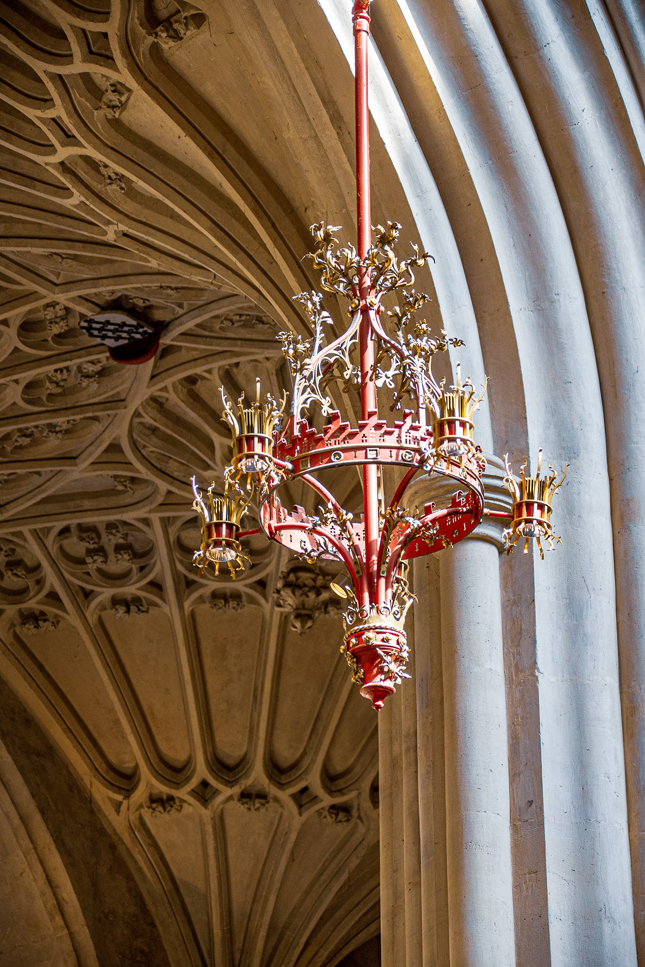 The image depicts an ornate, red and gold chandelier hanging from a ribbed, vaulted ceiling in what appears to be a cathedral or church. The ceiling features intricate stone carvings and ribbed arches, typical of Gothic architecture. The chandelier is elaborately designed with multiple branches and candle-like lights, adding to the grandeur of the setting.