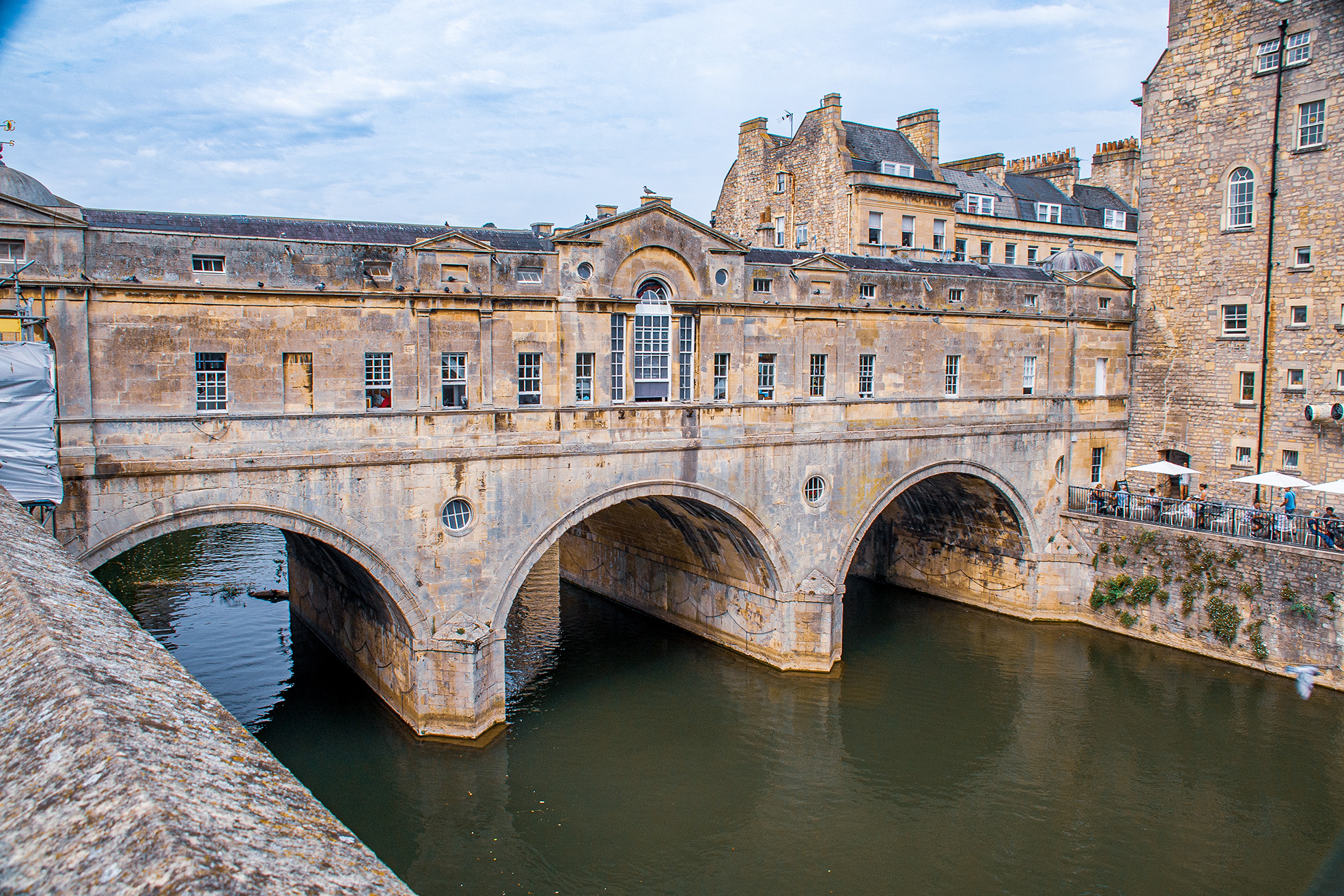 The image depicts the iconic Pulteney Bridge in Bath, England. This historic bridge spans the River Avon and is notable for the shops built into it, creating a unique architectural feature. The bridge is constructed of stone and features three wide arches. The buildings on the bridge have a classic Georgian architectural style, with large windows and chimneys. The scene is picturesque, with the river flowing calmly beneath the bridge and people visible enjoying the area.