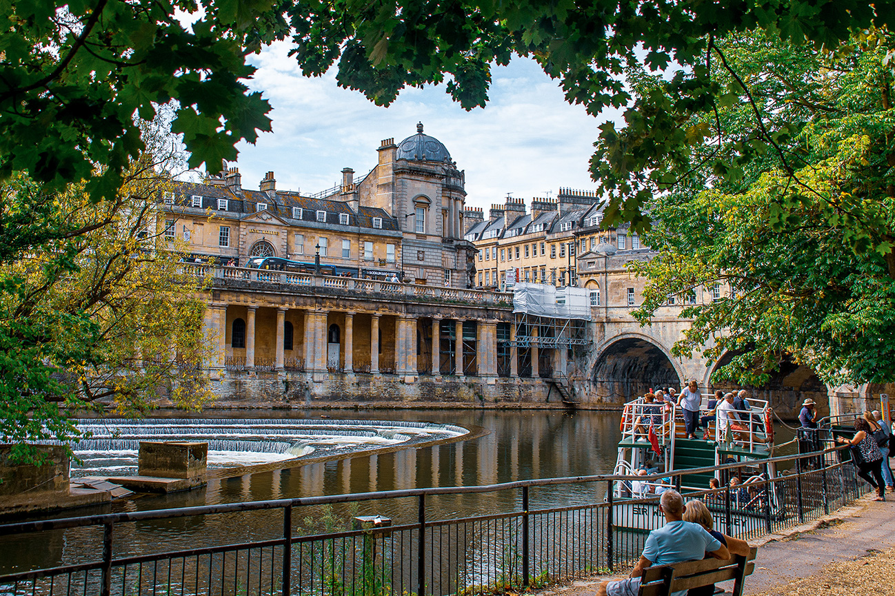 The image depicts the historic Pulteney Bridge in Bath, England. The bridge, which is unique for having shops across its full span, spans the River Avon. The architecture is classic Georgian style, with a large building atop the bridge. People are seen walking along the bridge and on a nearby boat. The scene is framed by lush green trees, adding a natural element to the urban setting.
