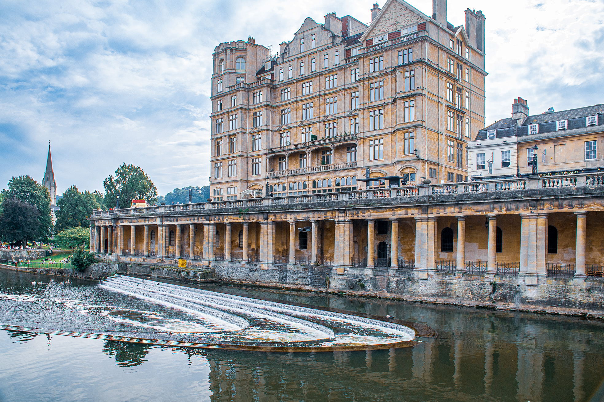 The image depicts the historic Bath Spa in Bath, England. The architecture features classic Georgian style with a multi-story building and a colonnade along the water's edge. The building is constructed with light-colored stone, and the scene includes a serene body of water with gentle cascades in the foreground. The sky is partly cloudy, and there are trees and other structures visible in the background, contributing to the picturesque setting.