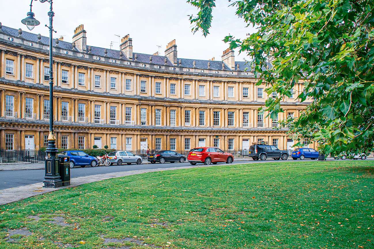 The image depicts a row of Georgian-style townhouses, characterized by their uniform architecture and symmetrical windows. The buildings are made of light-colored stone and feature multiple chimneys on the roof. In front of the townhouses, several cars are parked along the street, and a few motorcycles are also visible. The scene includes a well-maintained grassy area with a path and a lamppost, suggesting a quiet, residential neighborhood. The overall atmosphere is calm and picturesque, typical of a historic and well-preserved urban area.