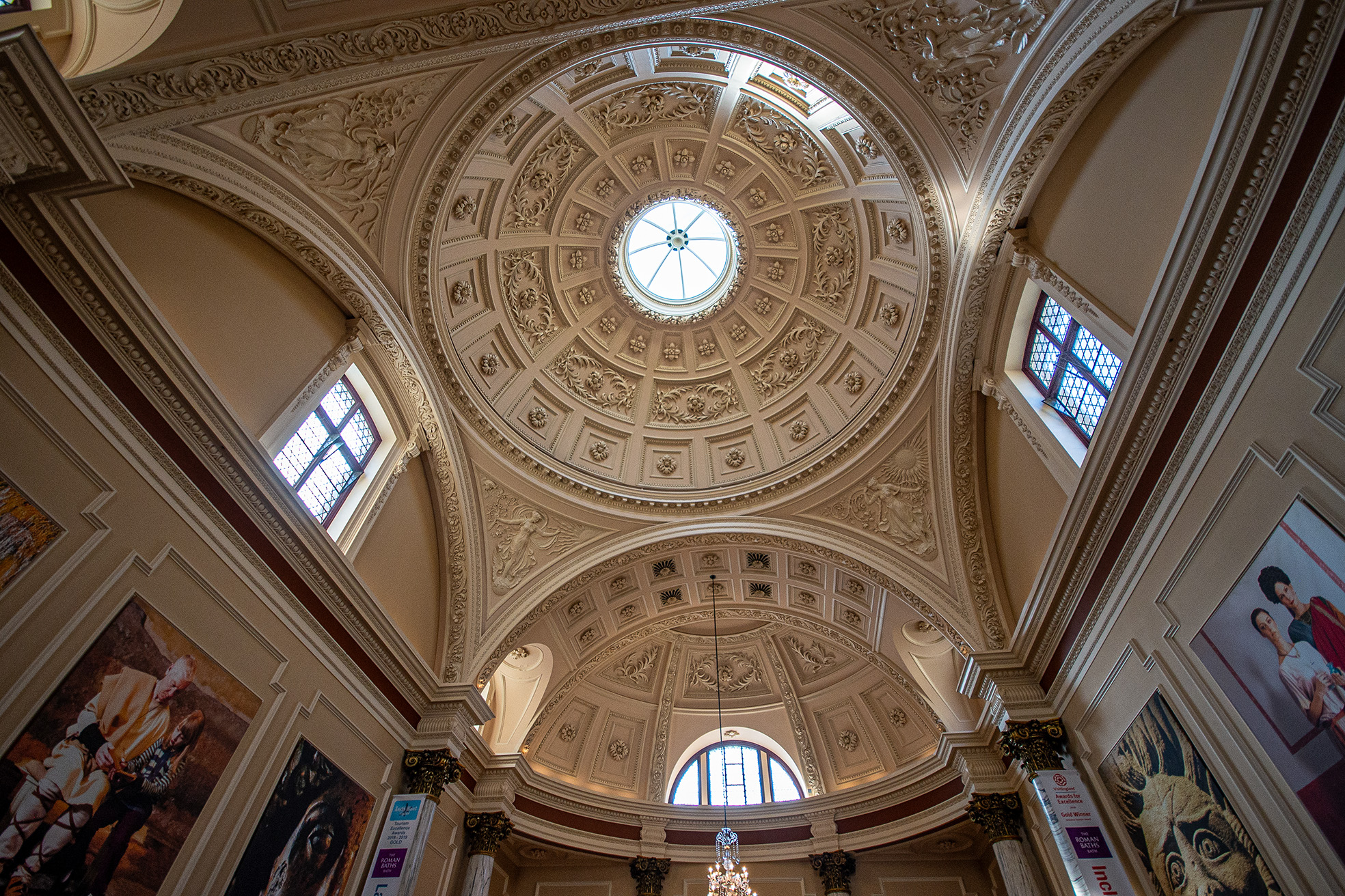 The image depicts the interior of a grand building, likely a museum or gallery, featuring an ornate, domed ceiling with intricate carvings and a central skylight. The walls are adorned with large paintings and decorative elements, and there are banners indicating exhibitions. The architectural style is classical, with detailed moldings and a sense of grandeur.