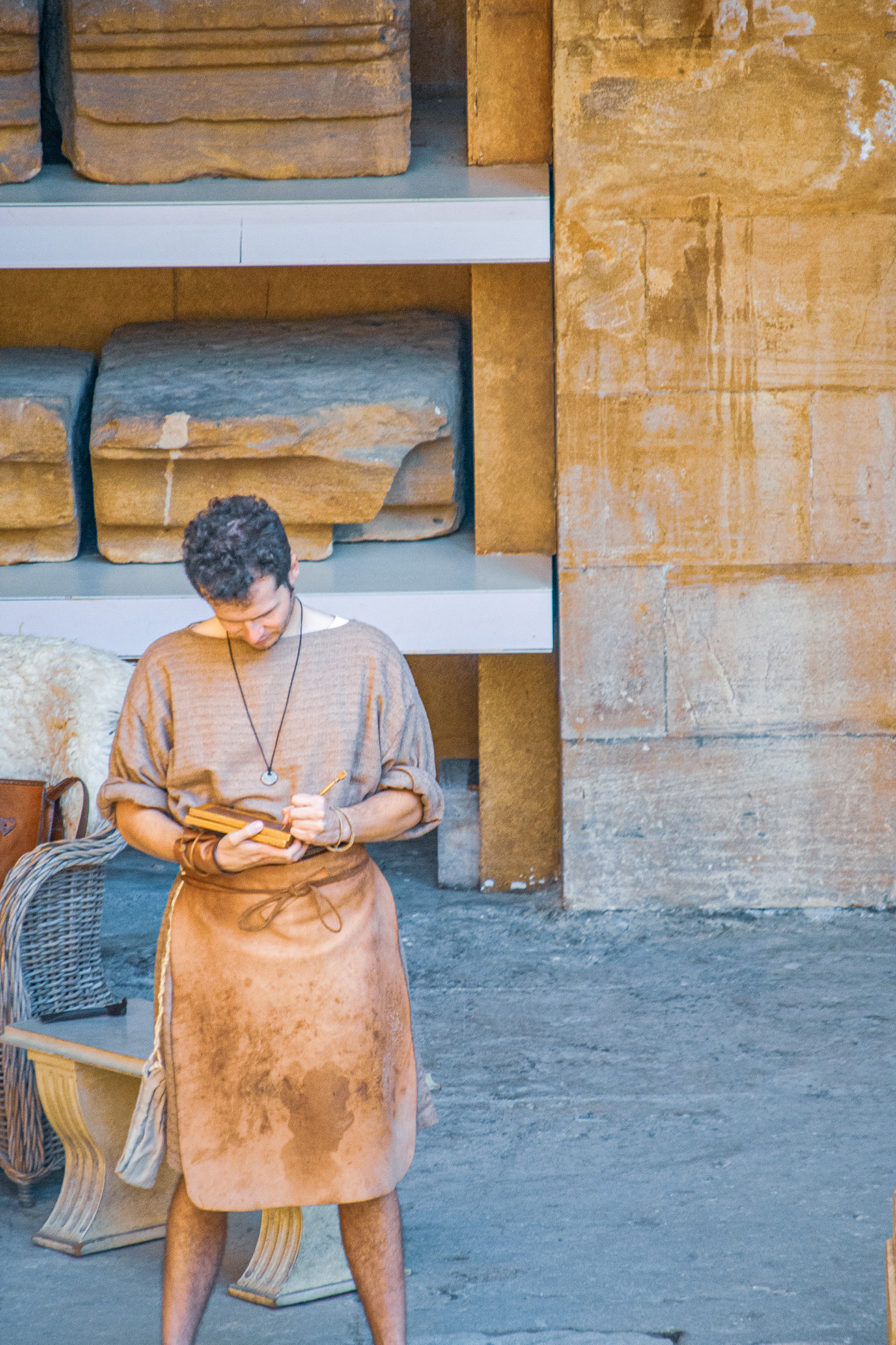The image depicts a person dressed in ancient attire, possibly a historical reenactor or actor, engrossed in reading a scroll. The setting appears to be a historical or archaeological site, with large stone blocks and structures in the background. The individual is wearing a beige tunic and a necklace, and is standing on a stone floor. The overall scene suggests a historical or educational context, possibly related to ancient civilizations or historical reenactments.