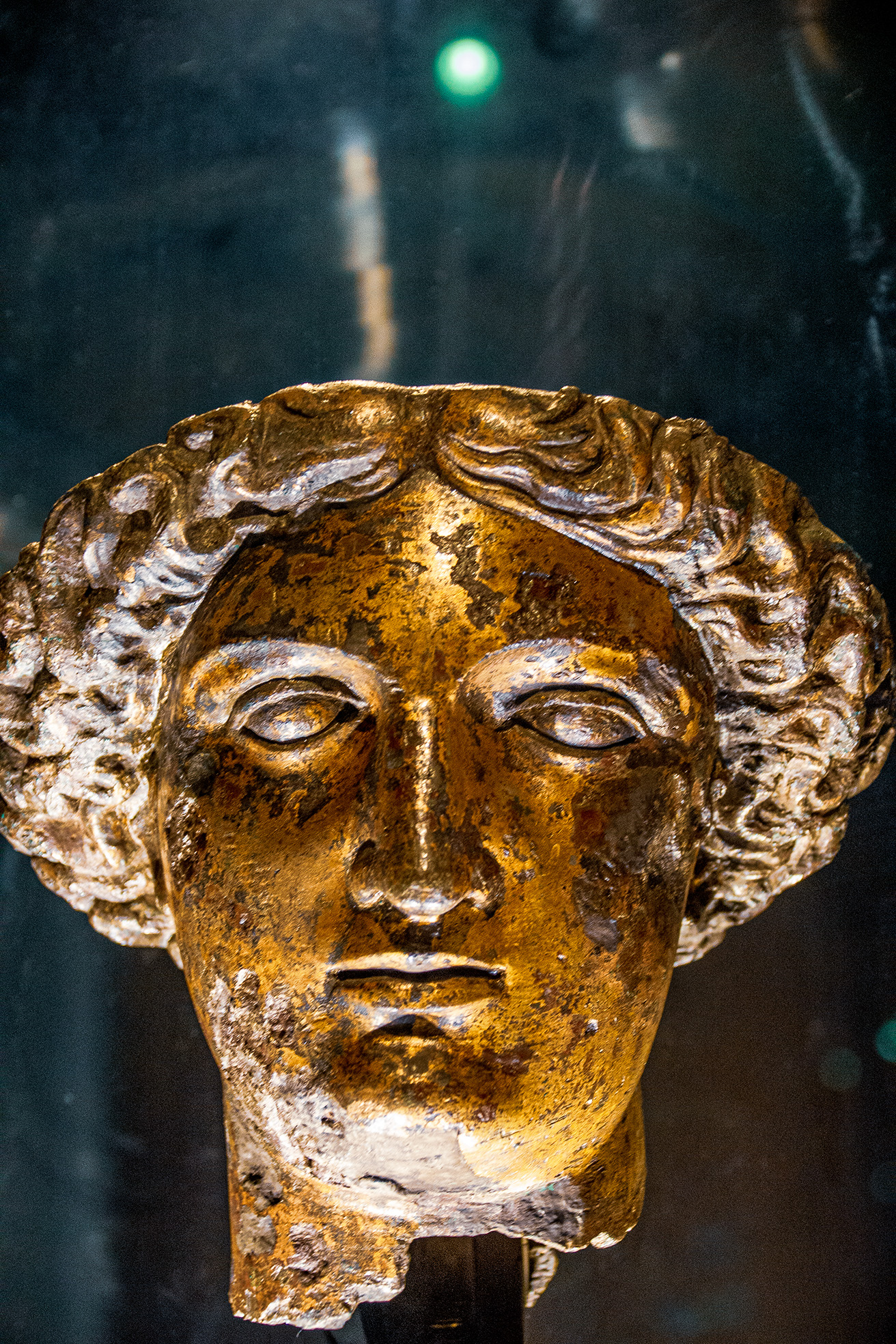 The image depicts an ancient golden artifact, specifically a mask, which appears to be made of metal. The mask has detailed features including wavy hair and pronounced facial characteristics. It is likely an artifact of historical or archaeological significance, possibly from ancient times. The mask is displayed against a dark background, highlighting its golden color and intricate design. The artifact is mounted on a stand, suggesting it is part of a museum or private collection.