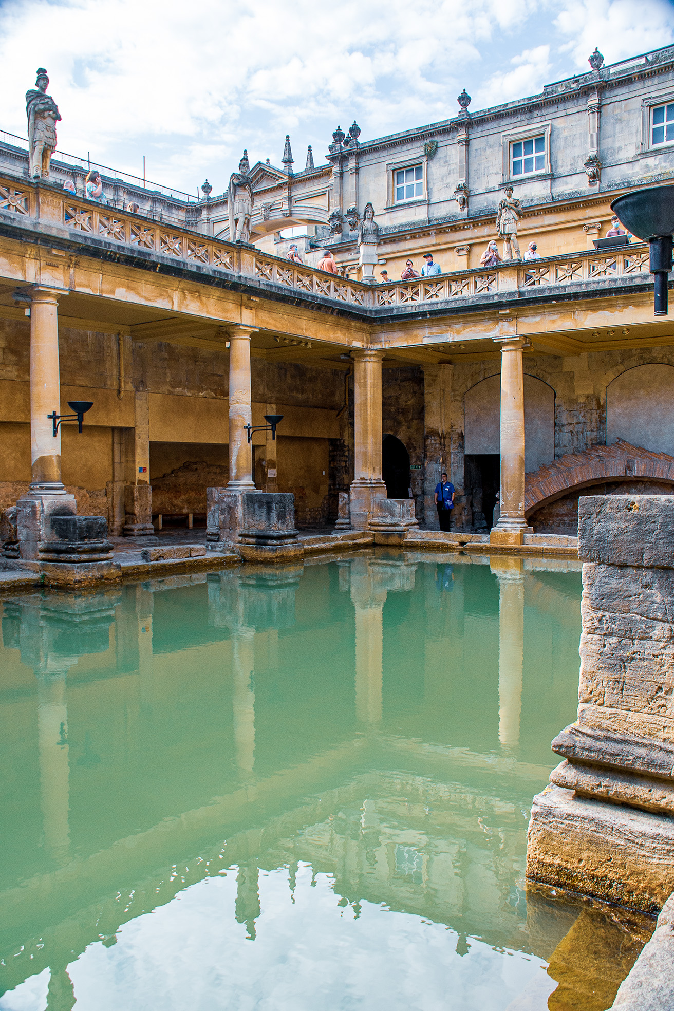 The image depicts the ancient Roman Baths in Bath, England. The scene shows a historic pool surrounded by classical architecture, including columns and statues. The water in the pool is greenish, reflecting the surrounding structures. There are people walking along the upper terrace, observing the site. The overall atmosphere is one of historical significance and architectural beauty.