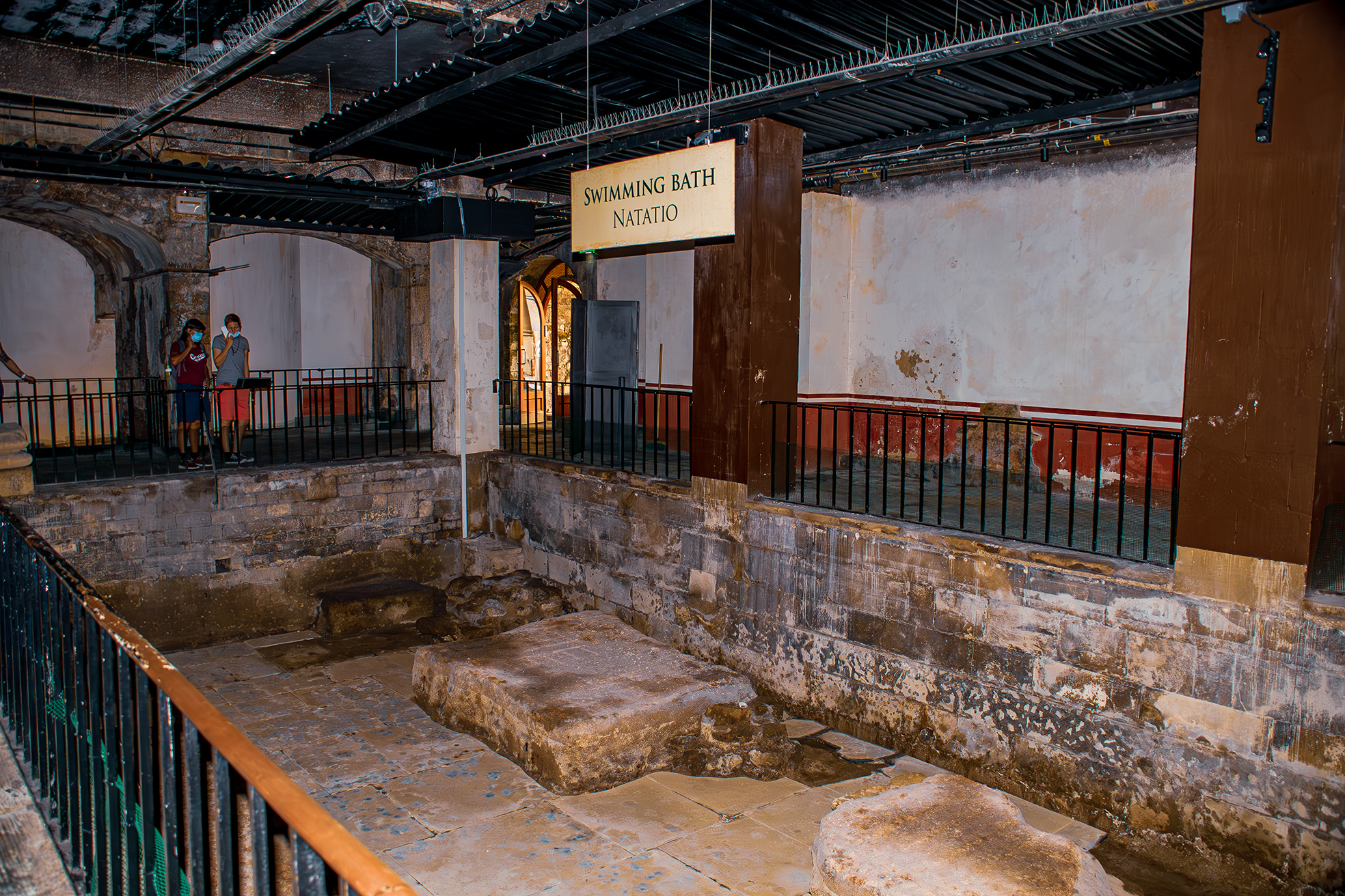 The image depicts an ancient Roman swimming bath, identified by a sign reading 'SWIMMING BATH NATATIO.' The structure features worn, stone walls and steps leading into the bath area, which appears to be partially filled with water. Two individuals are seen observing the site from a raised walkway, which is enclosed by metal railings. The ceiling above the walkway is equipped with modern lighting, suggesting the site is preserved and possibly open to the public for viewing.
