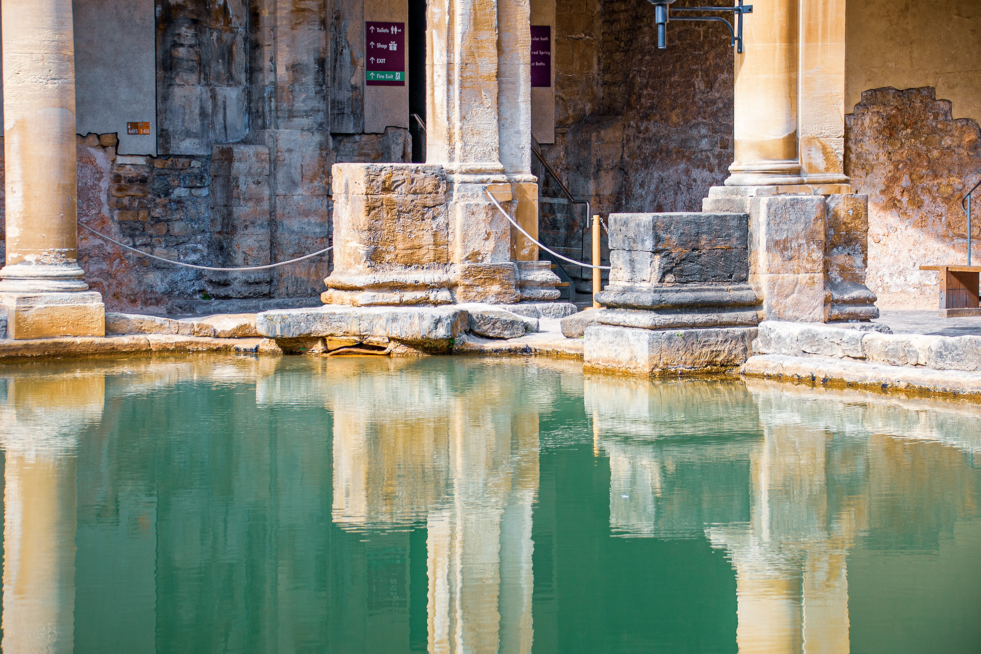 The image depicts an ancient Roman architectural site with a pool of water in the foreground. The structure features weathered stone columns and bases, reflecting the passage of time. The water in the pool is calm, mirroring the columns and parts of the structure. There are signs in the background indicating directions to various amenities such as toilets, shops, and exits. The overall scene suggests a historical or archaeological site, likely a Roman bath or similar structure.