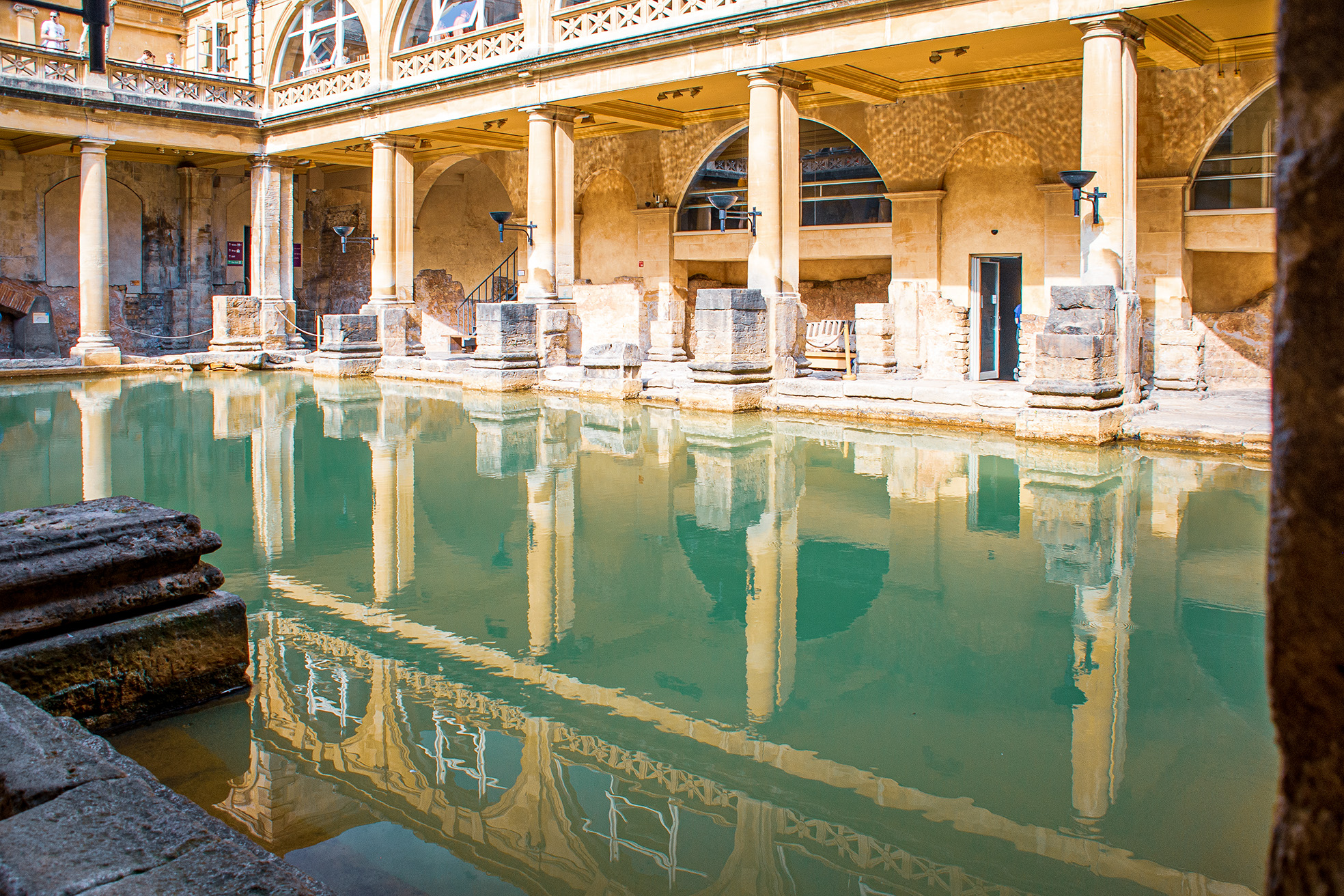 The image depicts the ancient Roman Baths in Bath, England. The scene shows a large, rectangular pool of greenish water surrounded by stone columns and arches. The architecture is classical, with a mix of well-preserved and weathered stonework. The upper level features intricate railings and large windows, while the lower level shows remnants of ancient stone structures. The overall atmosphere is serene, with the reflections in the water adding to the tranquility of the scene.