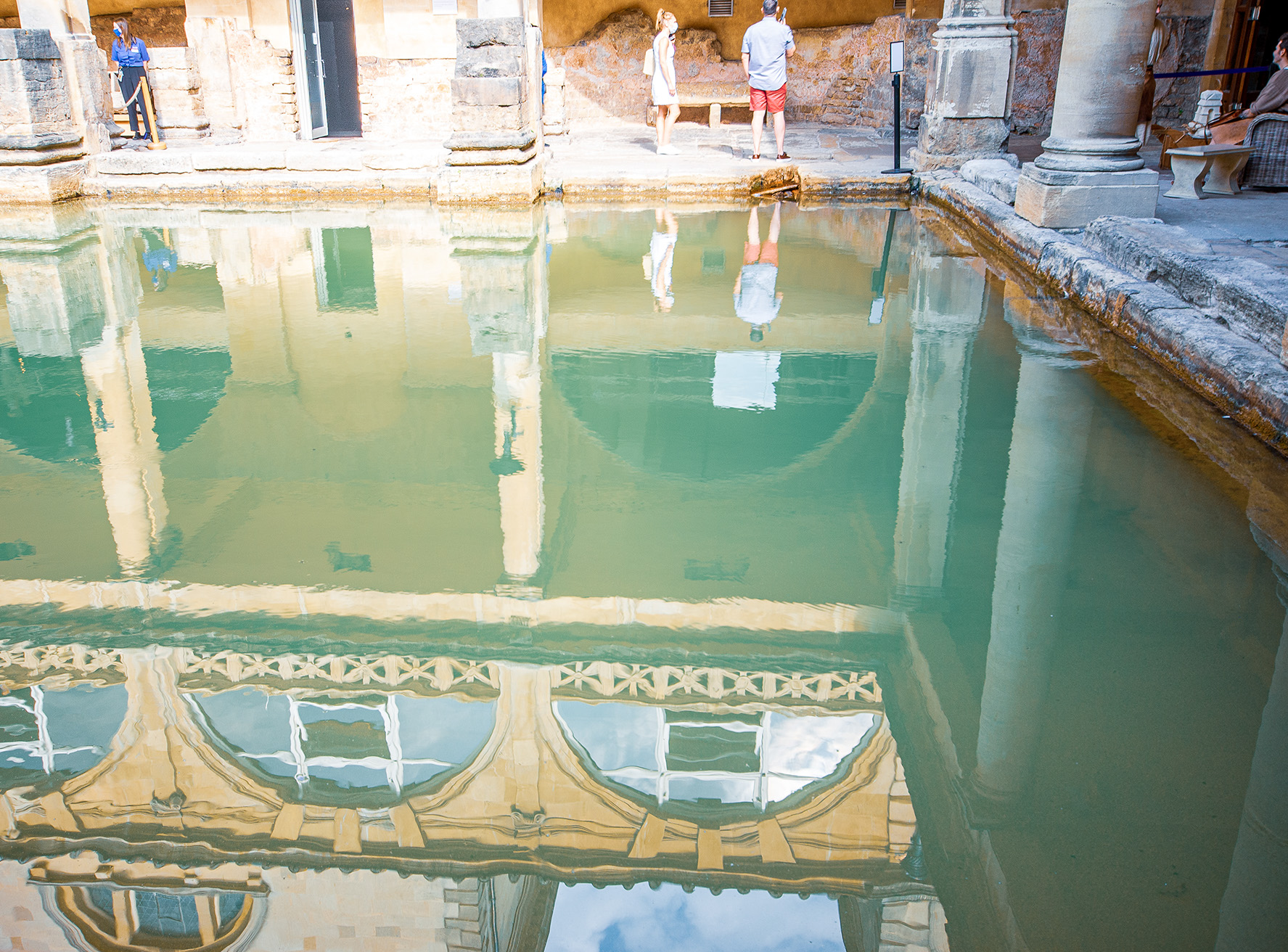 The image shows a historical Roman bath with a rectangular pool of greenish water, surrounded by stone columns and architectural details. Several people are standing and observing the bath, with their reflections visible in the water. The setting appears to be an ancient site, likely a tourist attraction, with stone walls and columns characteristic of Roman architecture.
