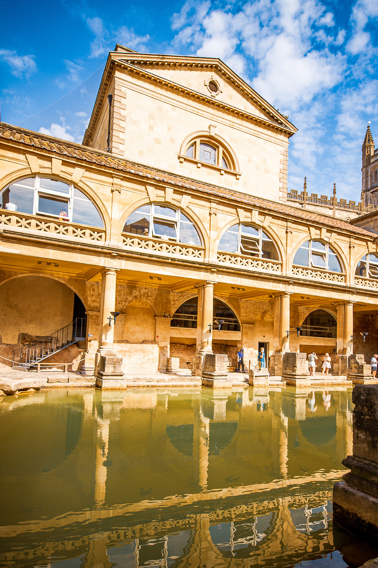 The image depicts the Roman Baths in Bath, England, showcasing the historic architecture and the ancient bathing pool. The structure features classic Roman design elements such as arches, columns, and intricate stonework. The pool reflects the building, creating a serene and picturesque scene. Visitors are seen walking around the site, indicating it is a popular tourist attraction. The sky is clear with a few clouds, adding to the overall bright and inviting atmosphere.