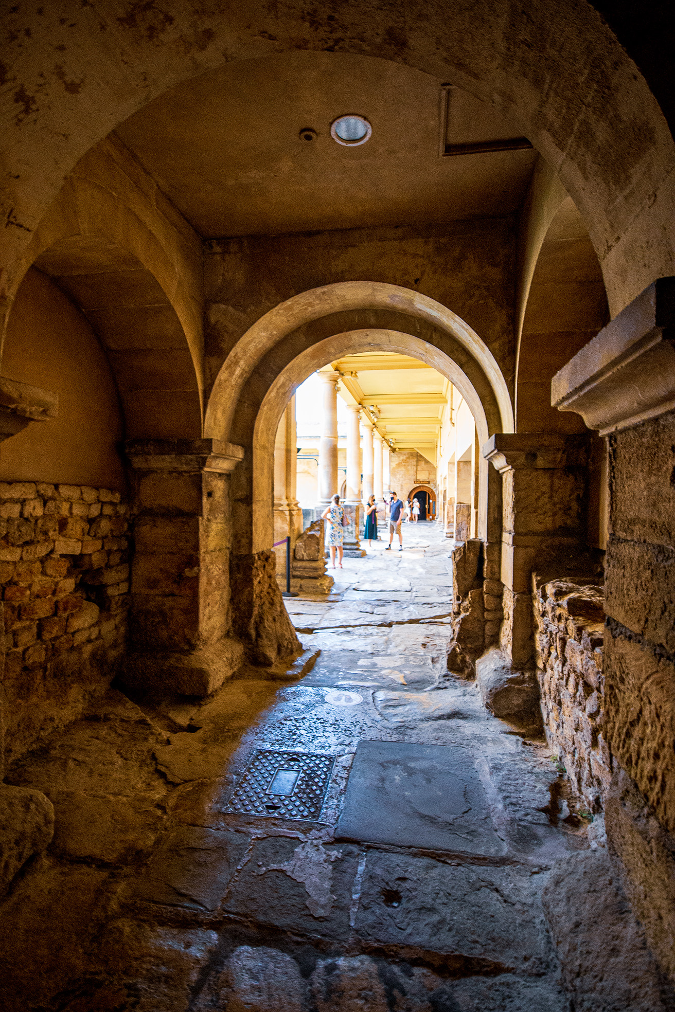 The image depicts a historical stone corridor with arched ceilings, leading to a courtyard with more arches and columns. The corridor is dimly lit, with light streaming in from the courtyard. The floor is made of stone slabs, and there are a few people walking and standing in the courtyard. The architecture suggests an ancient or medieval setting, possibly a historical site or ruin.