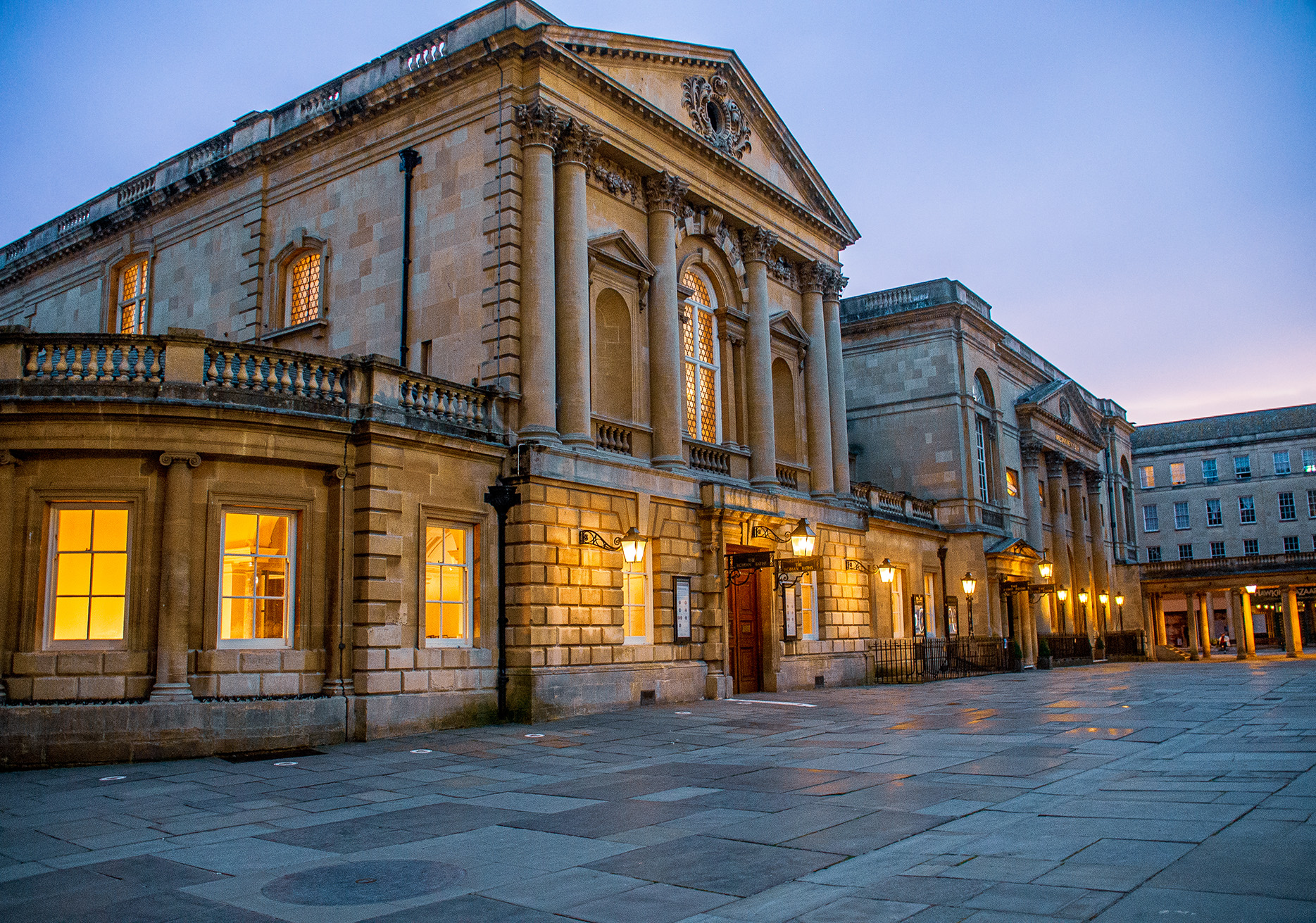 The image depicts a grand, historic building with classical architectural elements such as columns, pediments, and ornate detailing. The building is constructed from stone and features large windows that are illuminated from within, suggesting it is in use. The scene is captured during twilight, with a clear sky in the background. The structure is situated in a spacious, paved courtyard, and there are street lamps along the facade, adding to the elegant ambiance. The overall atmosphere is serene and majestic, indicative of an important or prestigious institution.
