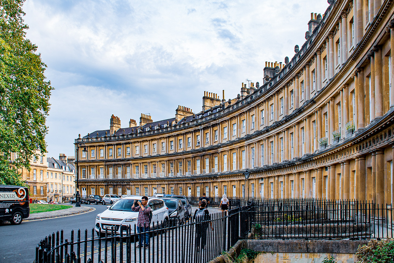 The image depicts a row of Georgian townhouses in a curved terrace formation, likely in Bath, England. The architecture features classic elements such as uniform facades, tall windows, and chimneys. The scene includes parked cars, a bus, and a few pedestrians. The sky is partly cloudy, and there is a tree on the left side of the image. The overall atmosphere is calm and picturesque.