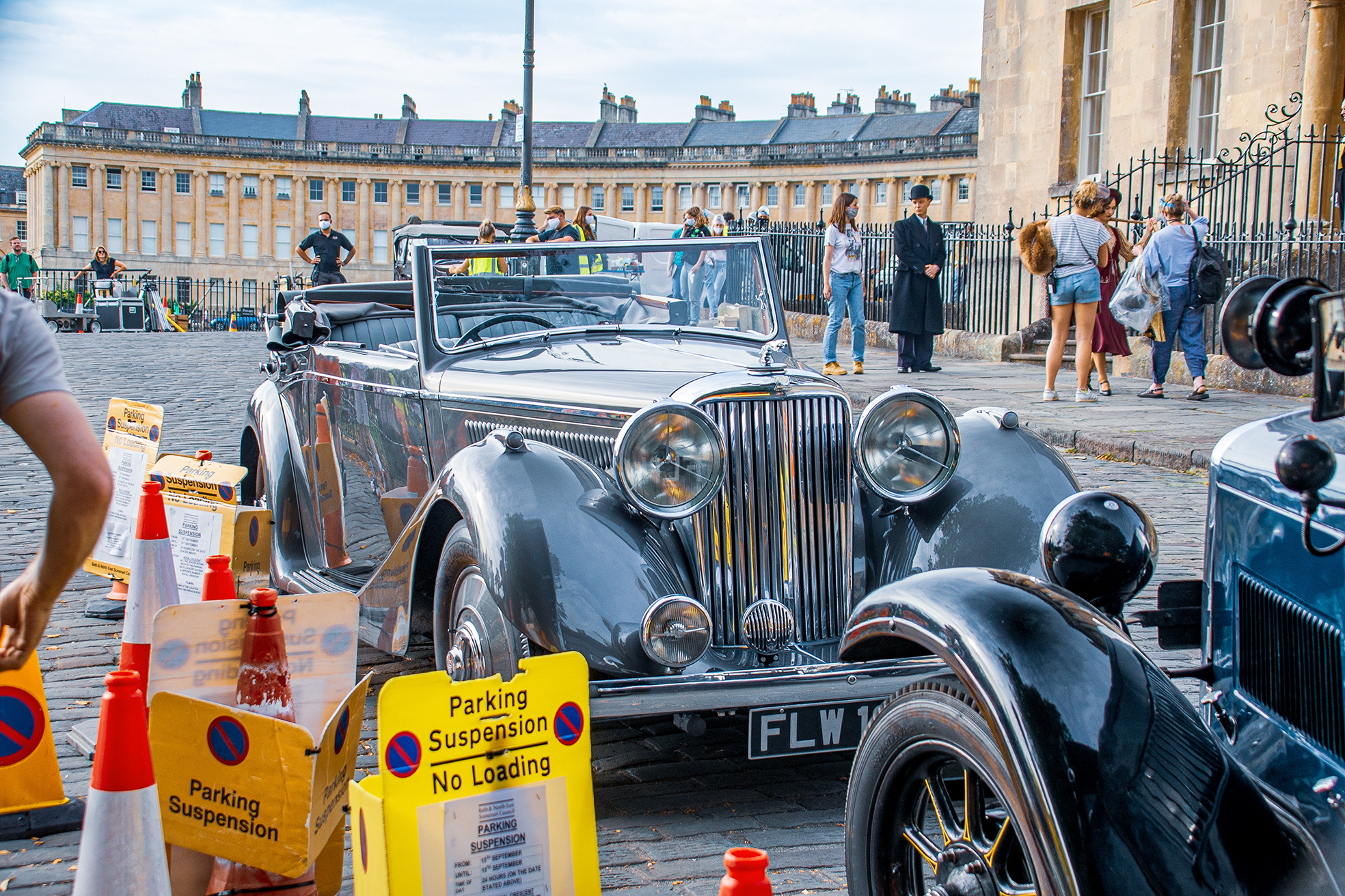 The image depicts a classic car parked in a historic square with a suspension parking sign nearby. People are gathered around, some taking photos, and the background features a large, elegant building with architectural details. The scene appears to be a public event or gathering, possibly a car show, with various individuals engaged in different activities.