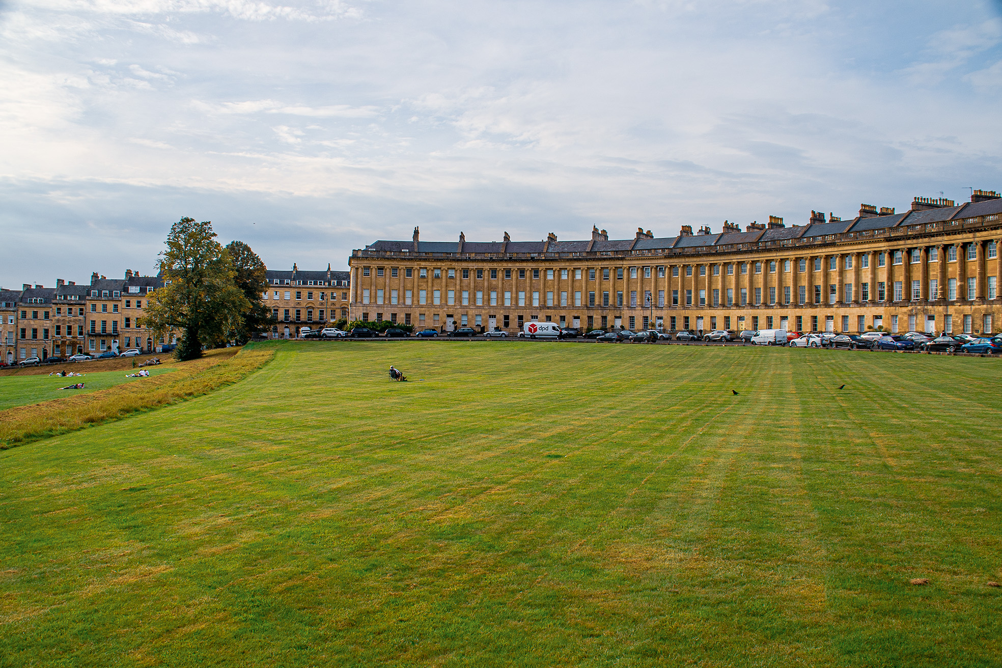 The image depicts a large, historic building with a symmetrical facade, featuring numerous windows and chimneys. In front of the building is a spacious, well-maintained grassy area with a few people and birds. The scene is set under a partly cloudy sky, and there are several parked cars along the street adjacent to the building.