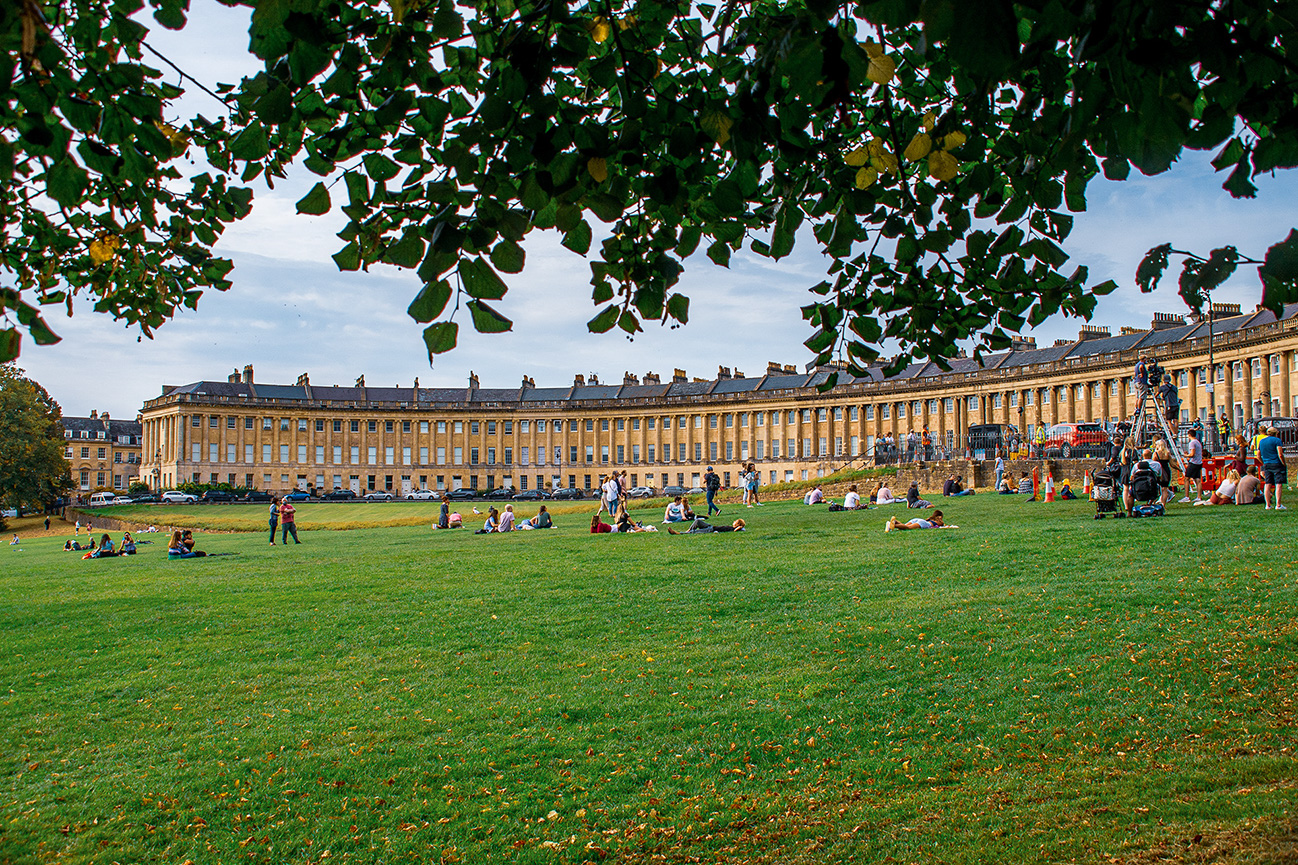 The image depicts a large, open green space with people relaxing and enjoying the outdoors. In the background, there is a long, historic building with classical architecture, featuring multiple windows and a uniform design. The scene is framed by tree branches at the top of the image, adding a natural border. The sky is partly cloudy, and there is some construction or filming equipment on the right side of the image.