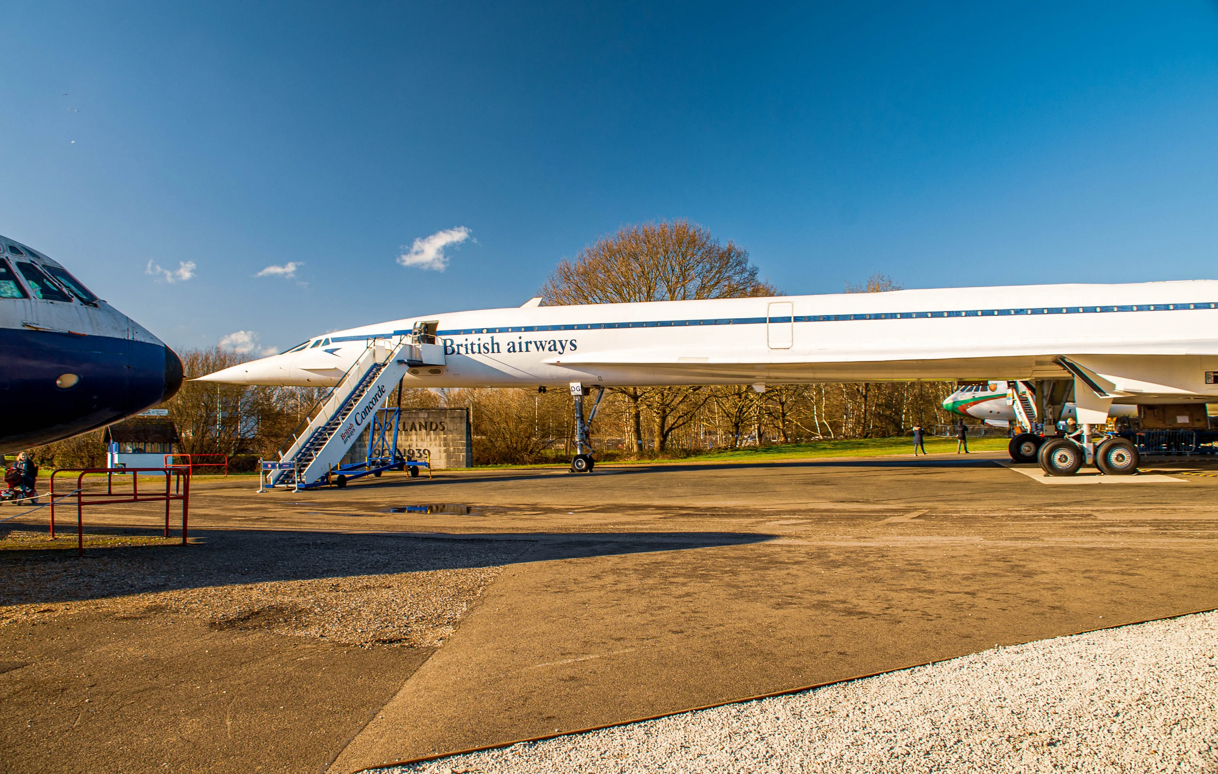 A British Airways Concorde aircraft is parked on display, with a mobile boarding staircase positioned beside it. The aircraft features the iconic white livery with blue stripes. The Brooklands Museum sign is visible in the foreground.