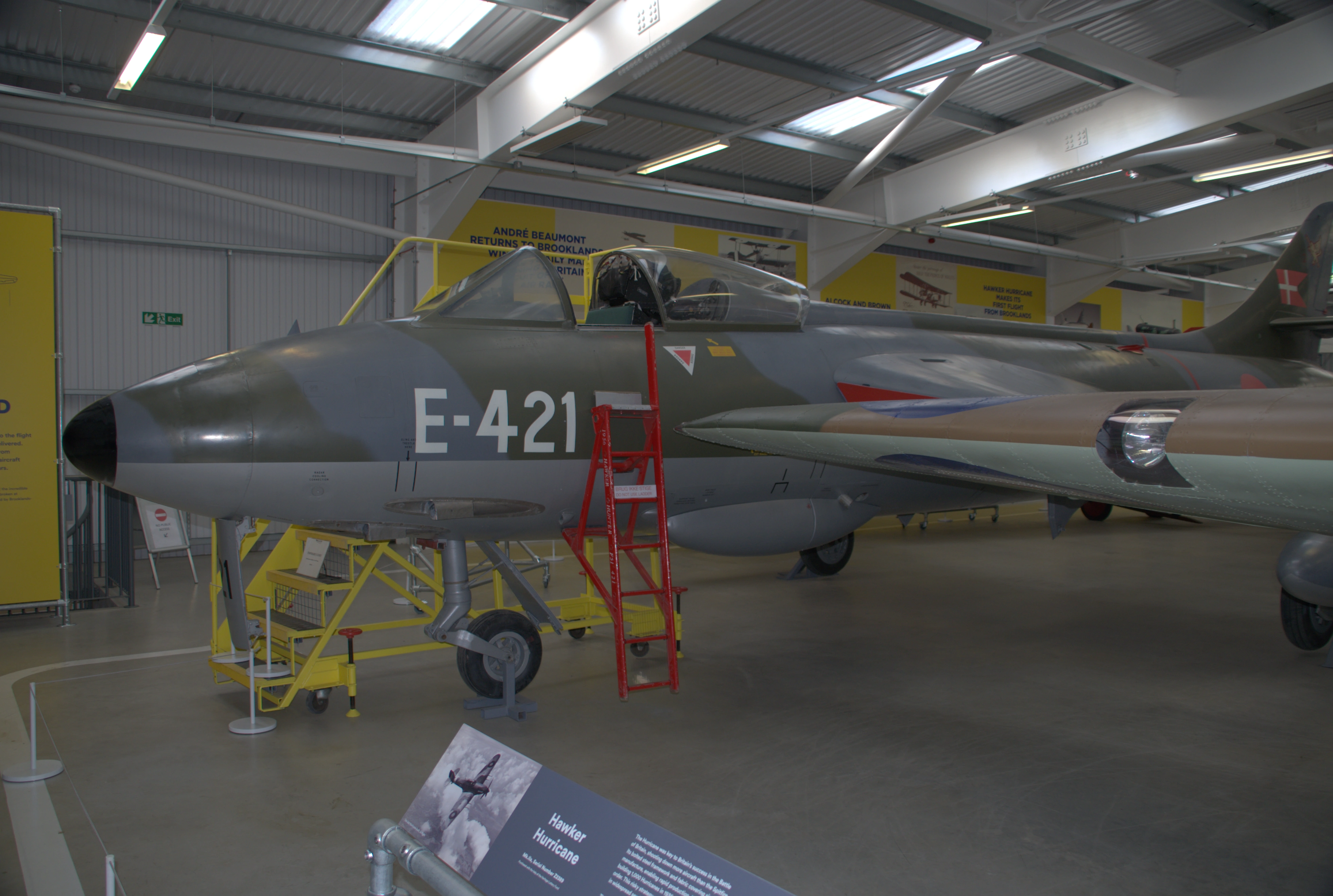 A museum hangar displays a collection of vintage aircraft, with a prominent dark blue jet in the foreground. A man stands to the right, viewing the aircraft.