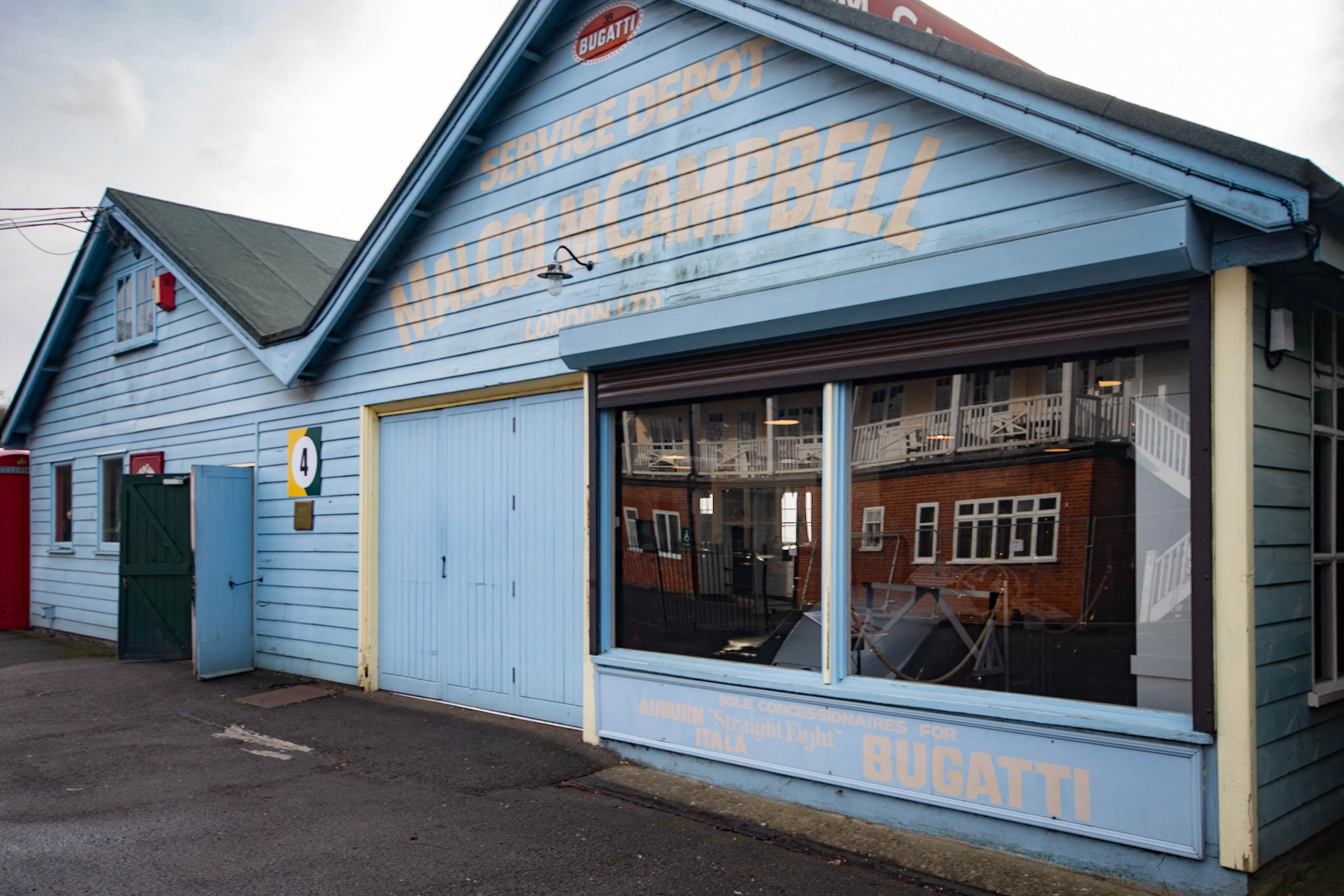 A light blue wooden building with cream lettering advertising Bugatti services and Malcolm Campbell London above a garage door and shop window, which reflects a brick building.