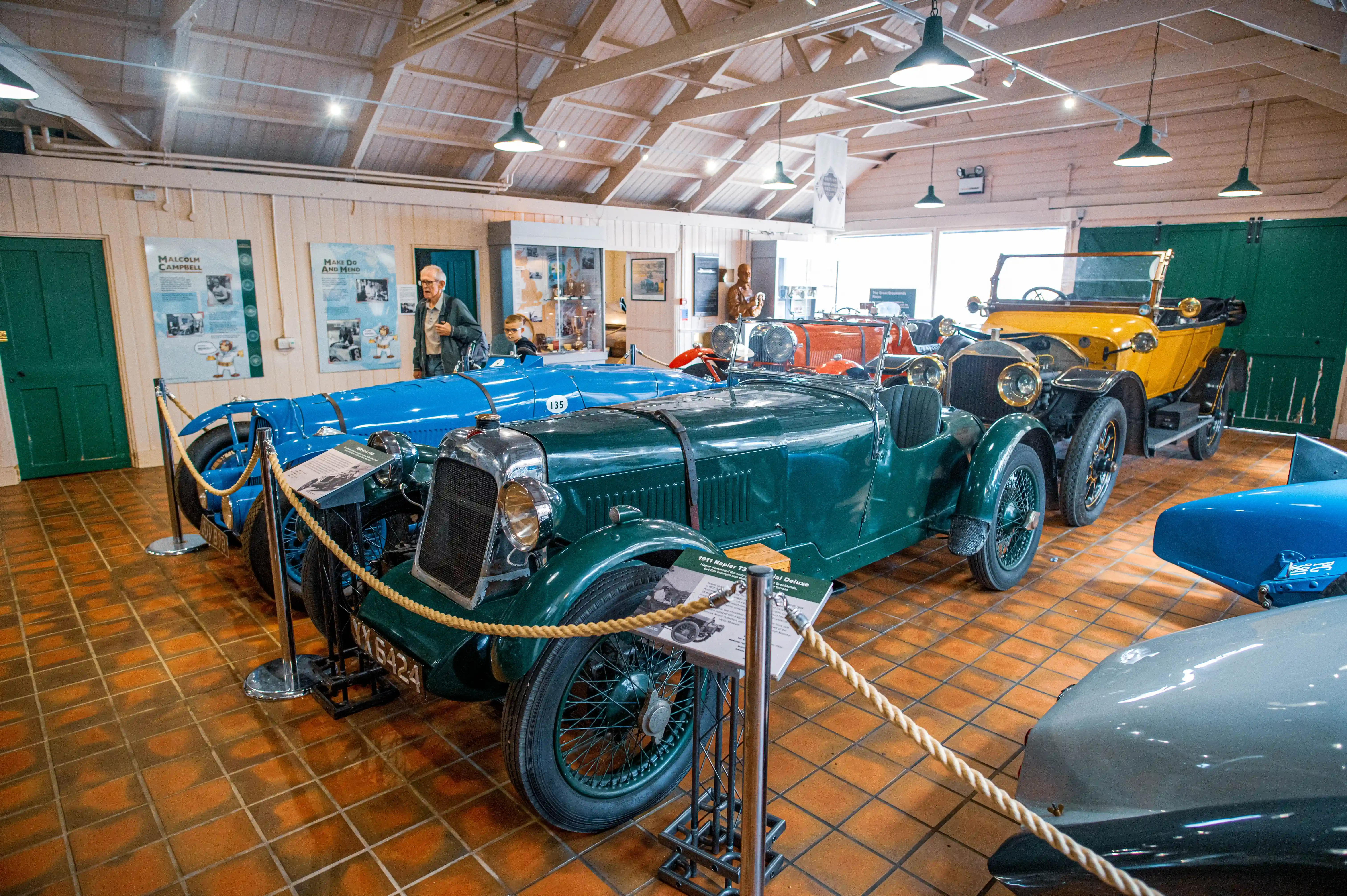 A museum interior showing several vintage cars in various colours, including green, blue, and yellow. Two visitors, an adult man and a child, are looking at the cars.