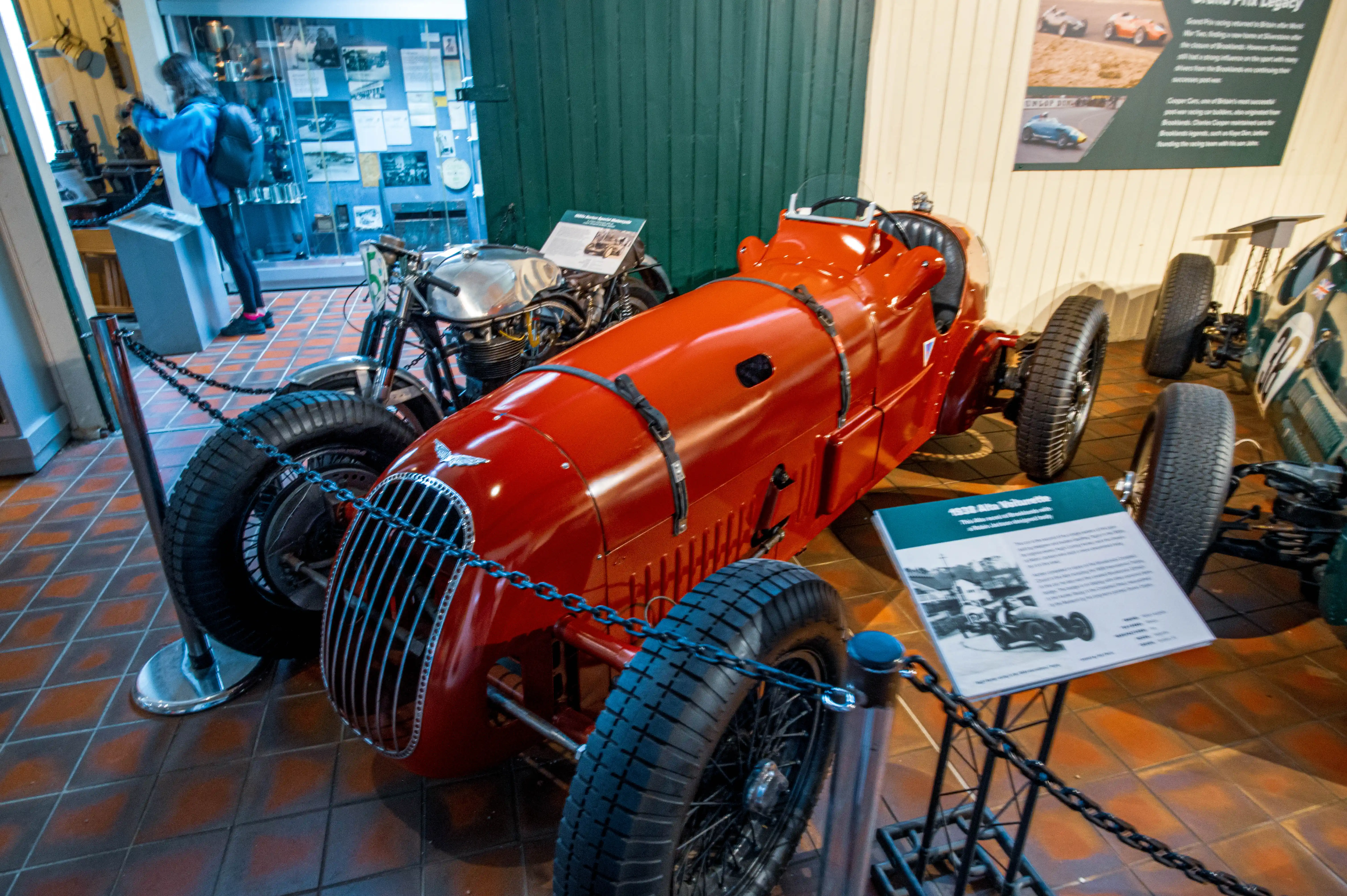 A bright red vintage racing car with a prominent grille and wire-spoked wheels sits on a tiled floor in a museum. A silver motorcycle is behind it, and a green racing car is to the right. A visitor in a blue jacket is in the background.