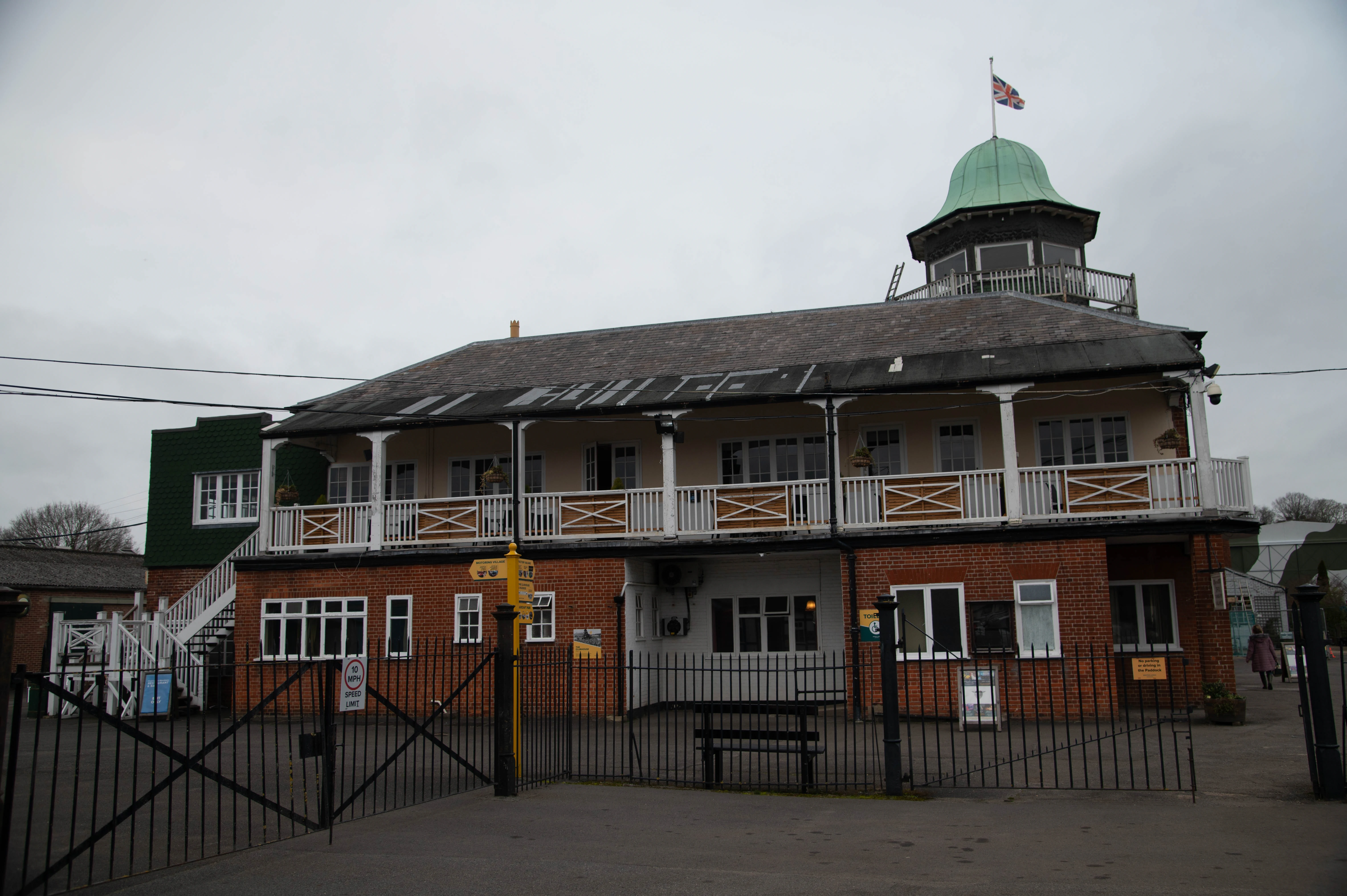 Exterior view of a two-story brick building, likely a cricket pavilion, with a prominent green-domed tower at the top. The tower flies a British flag. A balcony runs along the first floor, and a black metal gate is in the foreground.