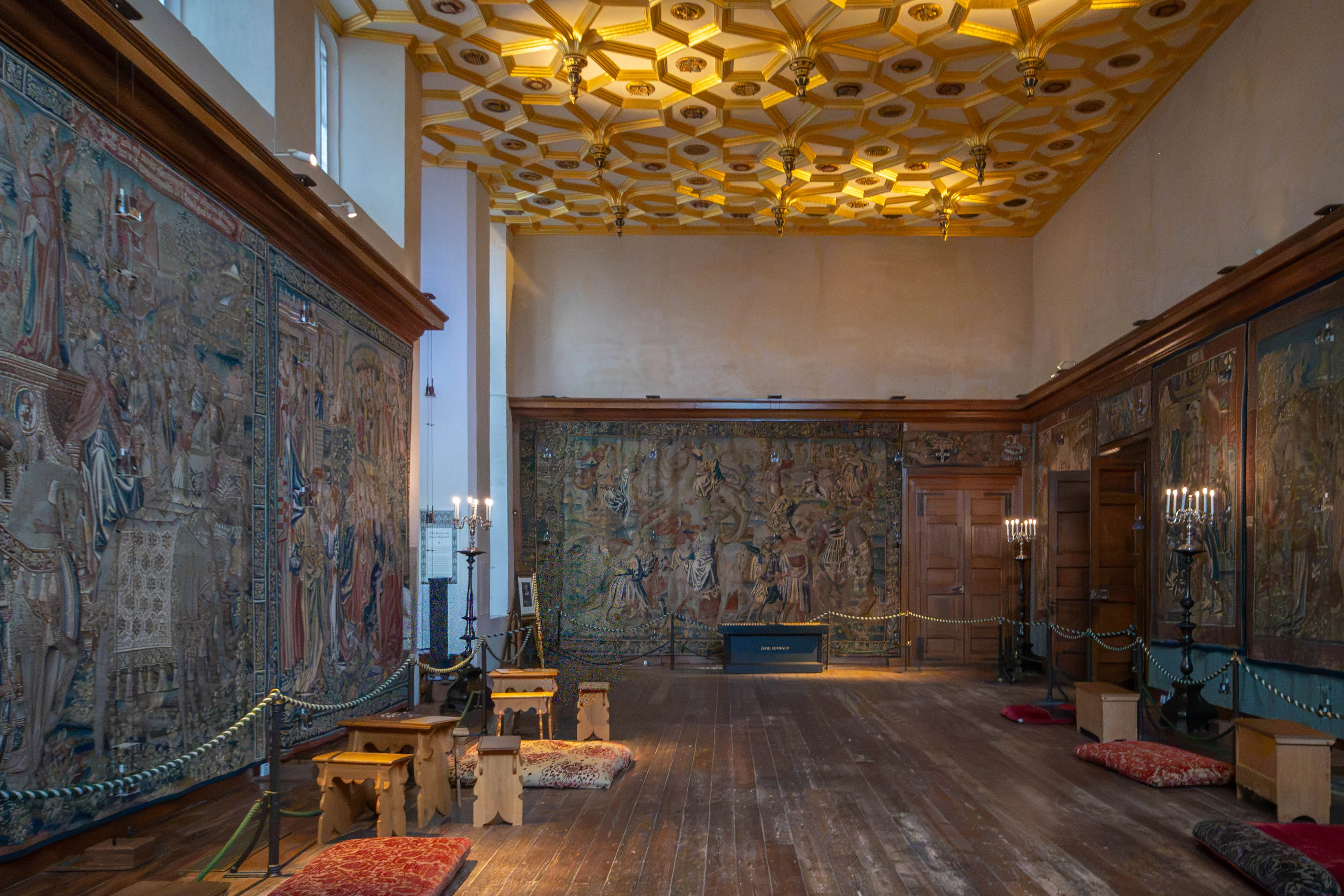 Historic room with large tapestries, gold-and-white geometric ceiling, cushioned benches and soft mixed lighting.