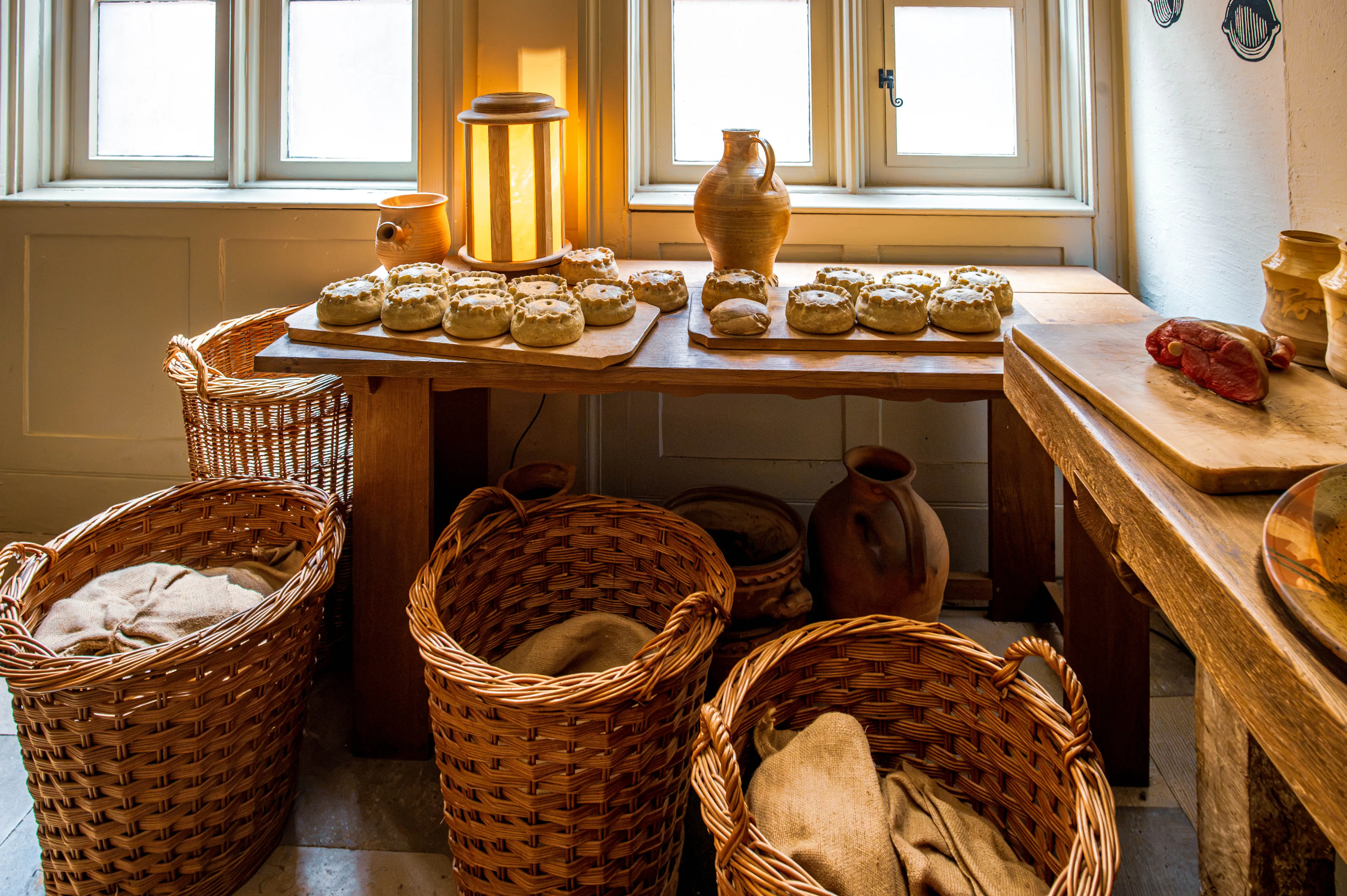 Rustic kitchen with wooden table, bread loaves, wicker baskets, ceramic jugs and warm window light.