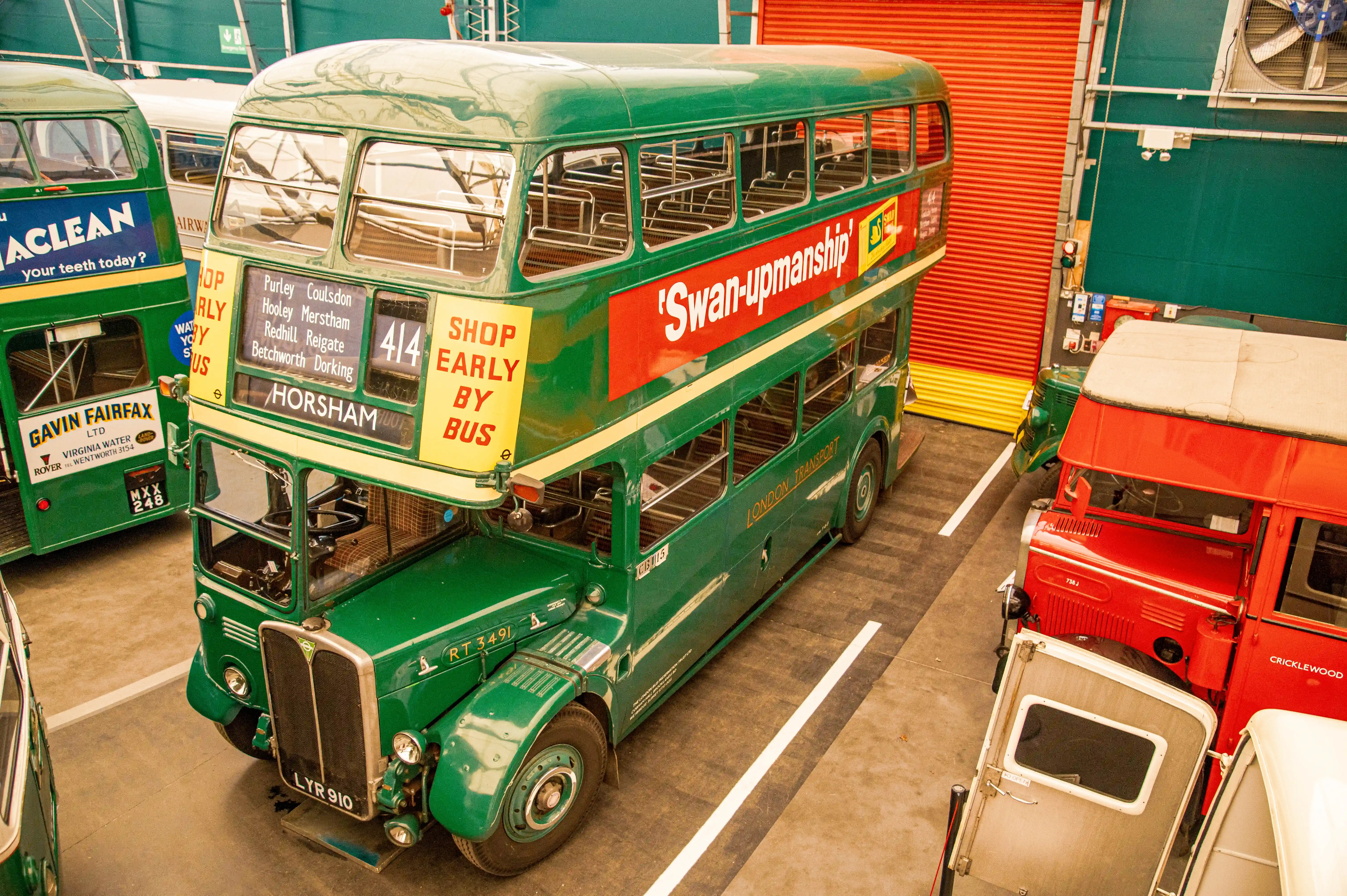 A vintage green double-decker London Transport bus with advertisements and route information is parked next to other vintage buses, including a red one, inside a building with a red roller shutter door.