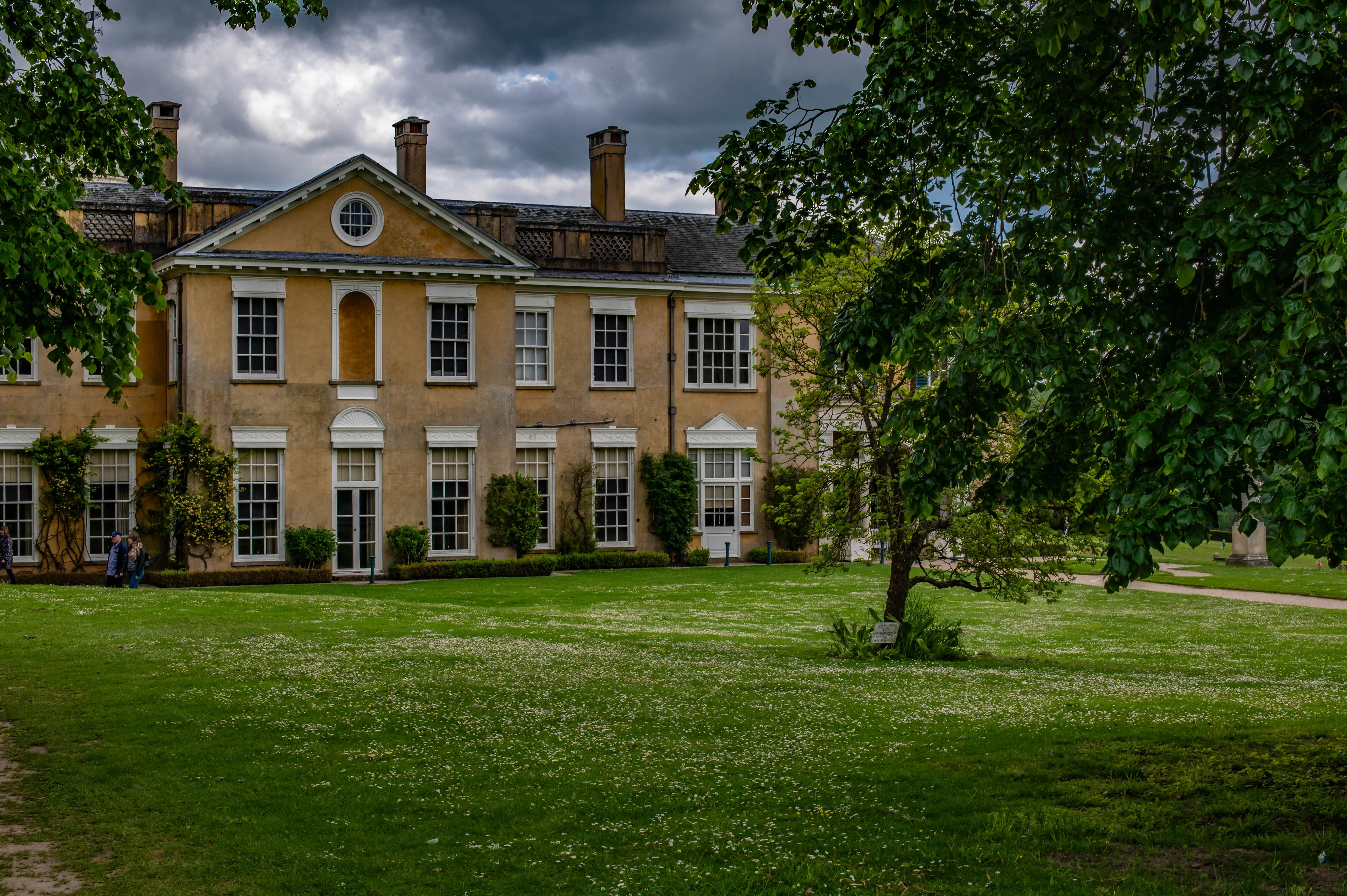 A large, pale yellow country house with white-framed windows and classical architectural details, viewed from a green lawn dotted with small white flowers under a dark, cloudy sky. Trees with lush green foliage frame the scene.