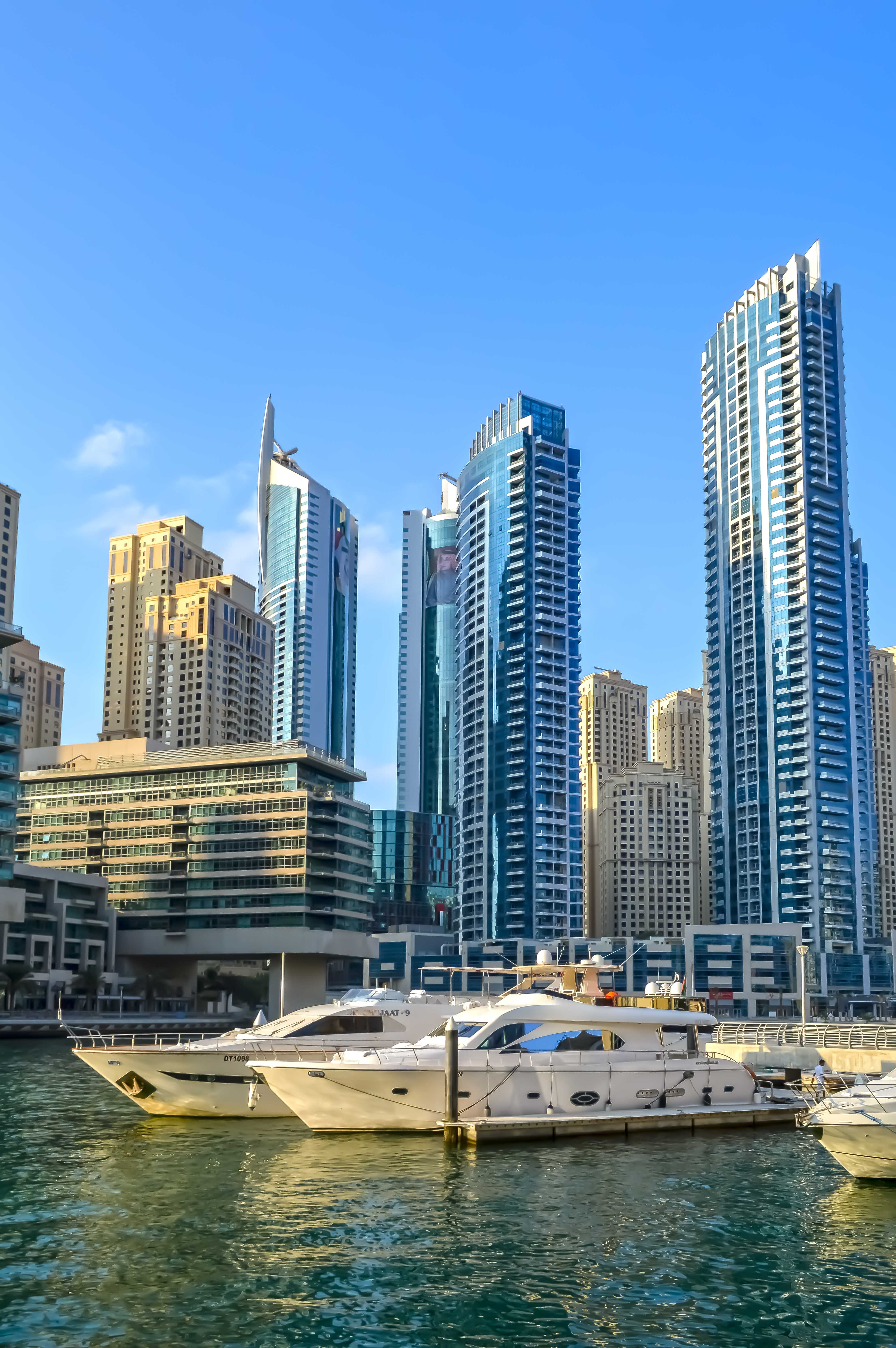 The image depicts a modern waterfront cityscape with tall, contemporary skyscrapers and a luxurious yacht docked in the foreground. The buildings are a mix of glass and steel, showcasing various architectural designs. The water is calm, reflecting the buildings and the yacht. The sky is clear, indicating a sunny day. The overall scene suggests a vibrant, urban environment, likely a major city known for its skyline and waterways.