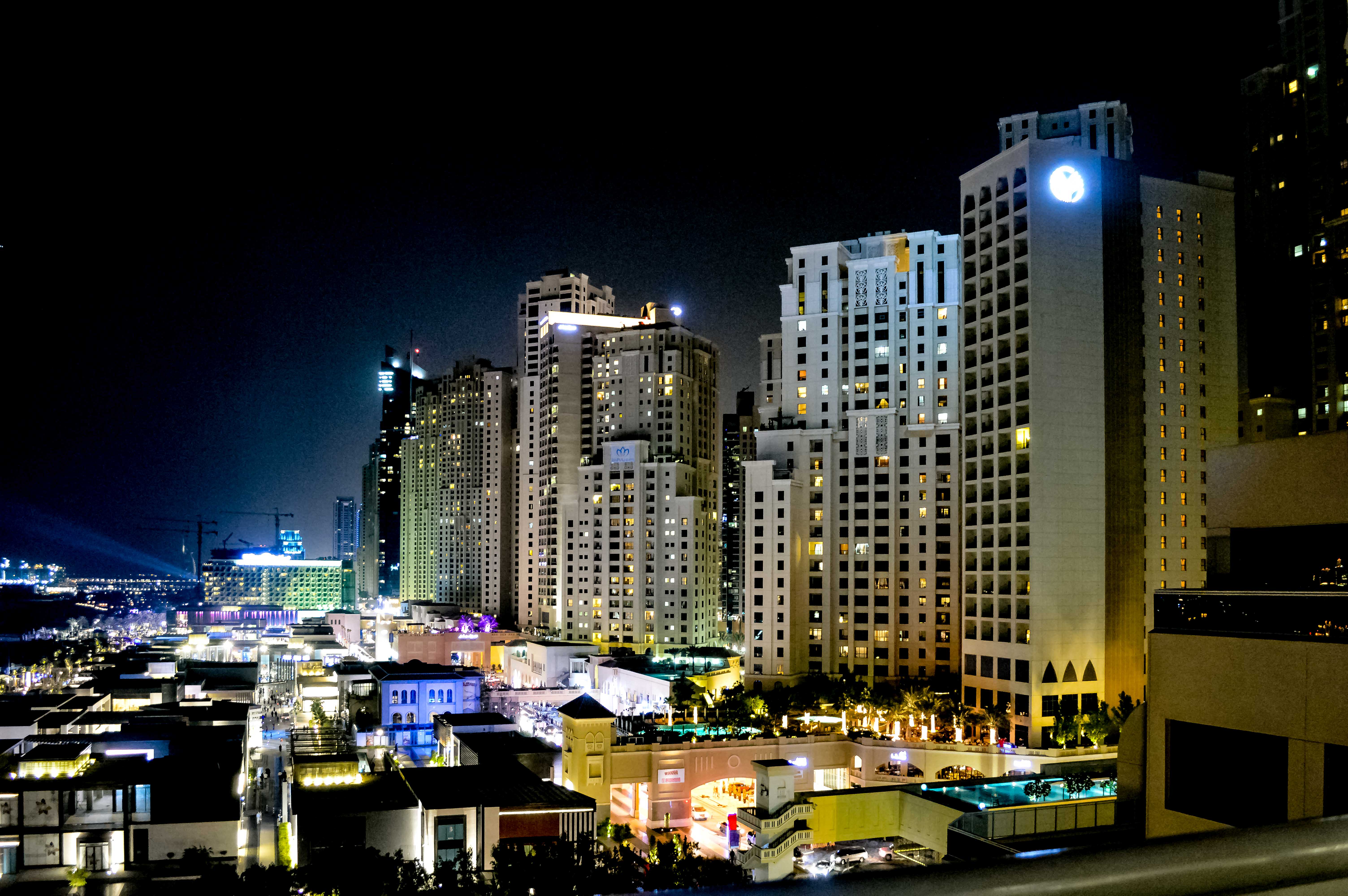The image depicts a vibrant cityscape at night, featuring a cluster of tall, illuminated buildings. The skyscrapers are adorned with numerous windows, many of which are lit, indicating activity within. The architecture is modern, with a mix of rectangular and slightly curved structures. The scene includes a variety of lights from street-level buildings and possibly some construction cranes in the distance, suggesting ongoing development. The overall atmosphere is lively and bustling, characteristic of a busy urban area.