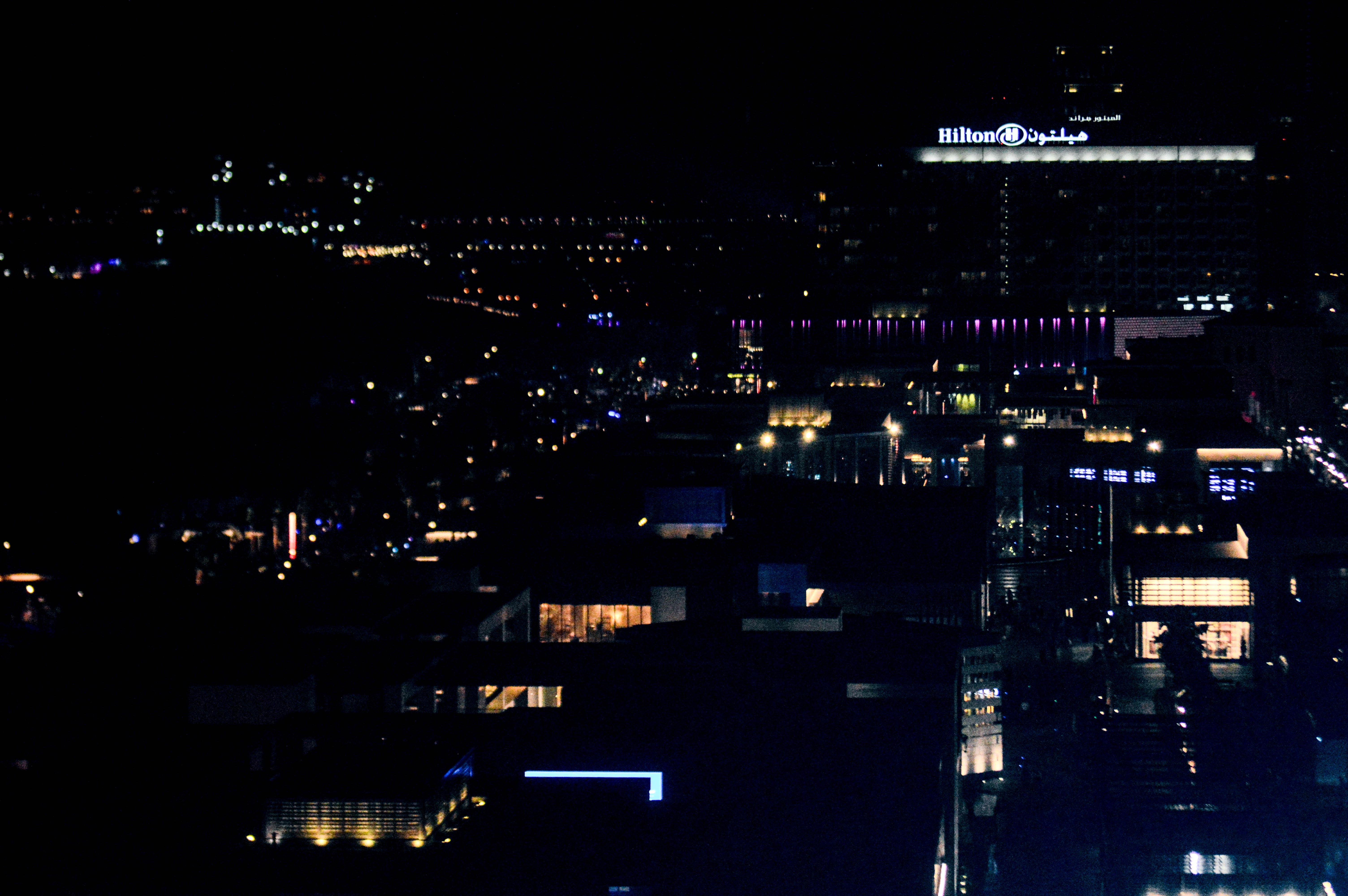 The image depicts a nighttime cityscape with illuminated buildings and streets. The most prominent feature is a large building with the Hilton logo lit up, indicating the presence of a Hilton hotel. The city appears to be bustling with lights from various buildings and streetlights, creating a vibrant urban scene.