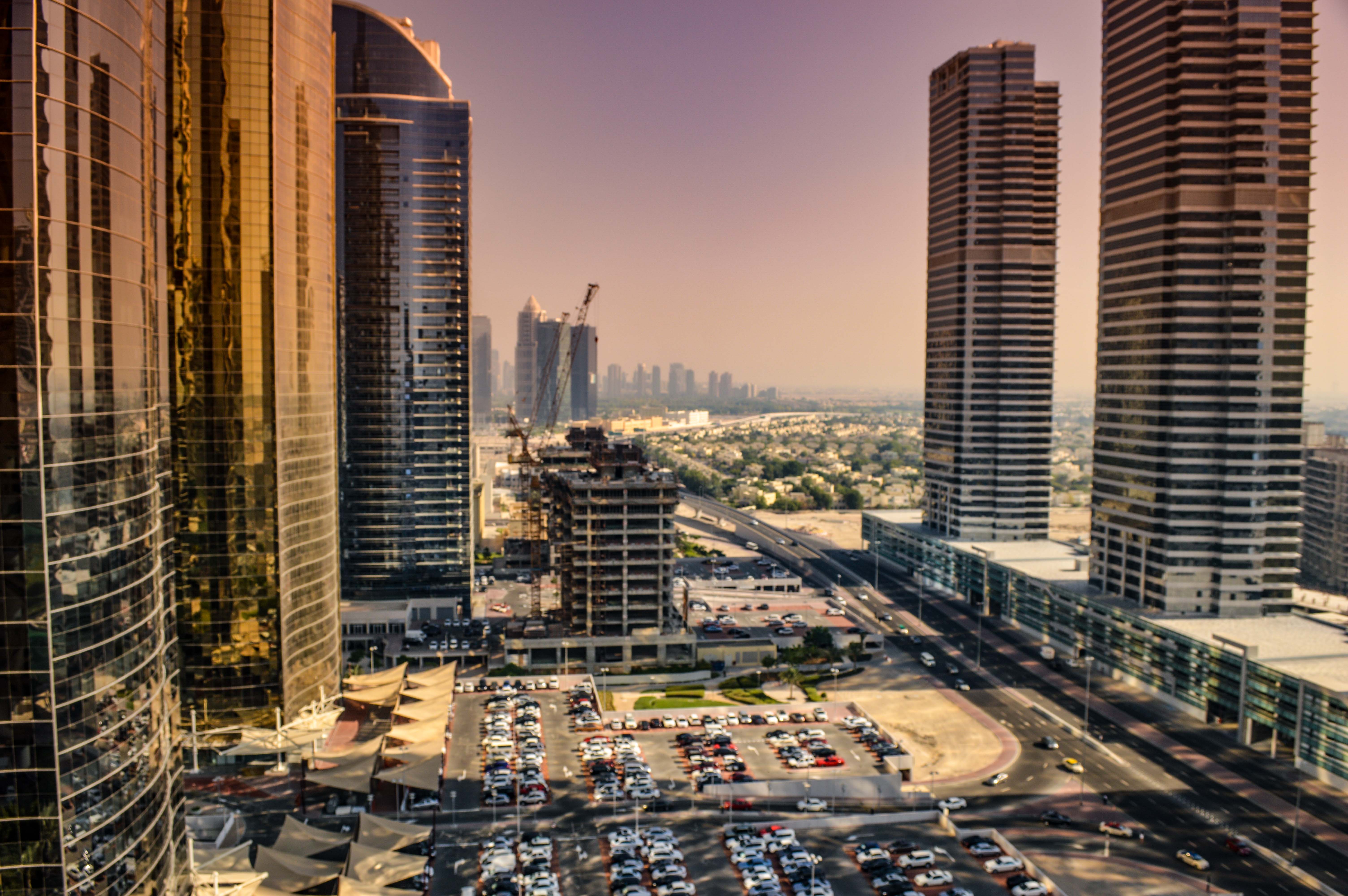 The image depicts a modern urban landscape with tall skyscrapers, construction cranes, and a busy street filled with cars. The scene is likely set in a bustling city, showcasing a mix of completed and under-construction buildings. The architecture is contemporary, with reflective glass facades and sleek designs. The area appears to be a commercial district, given the presence of parking lots and the density of the buildings. The overall atmosphere suggests a dynamic and rapidly developing cityscape.