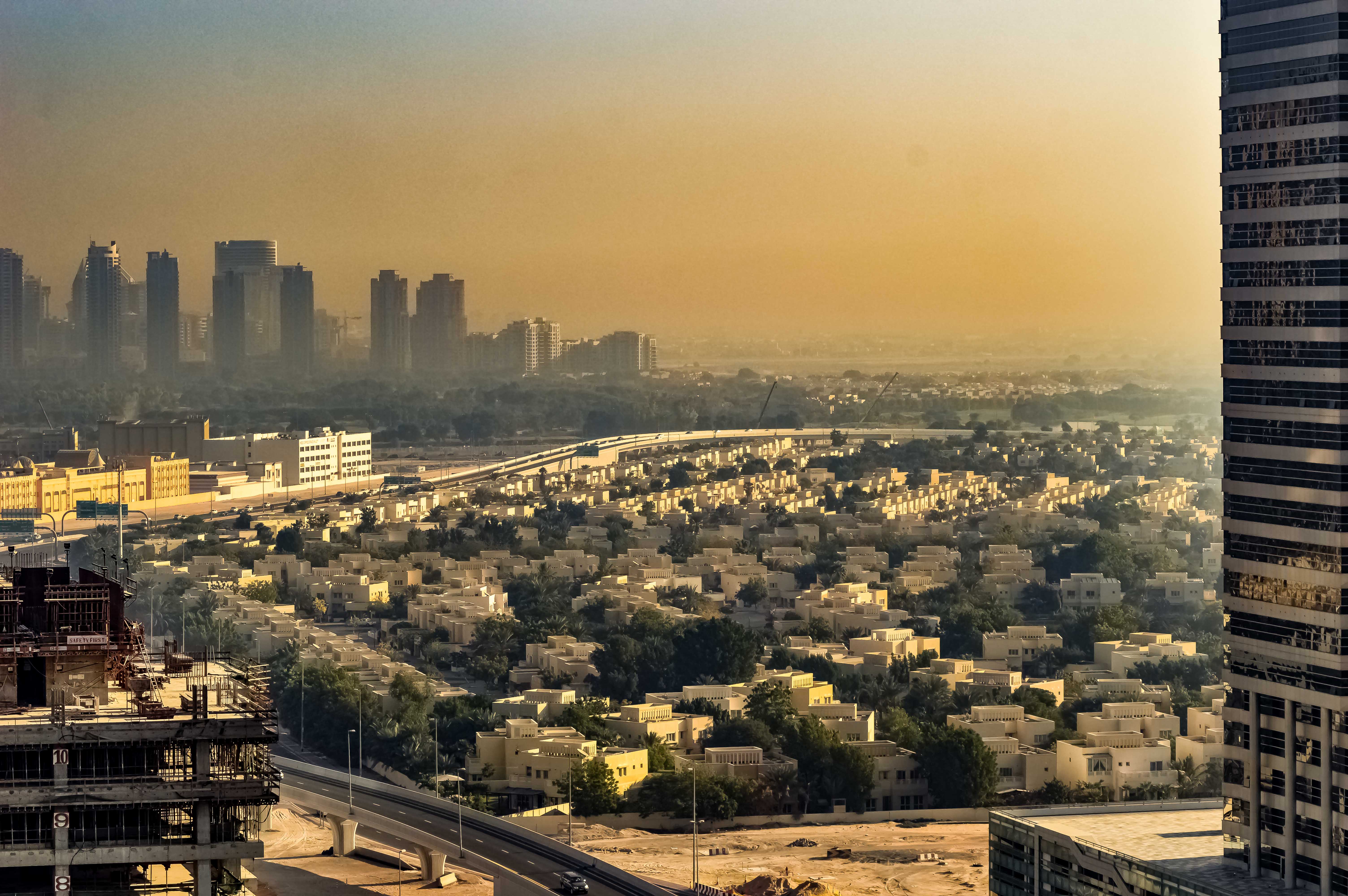 The image depicts an urban landscape with a mix of high-rise buildings and low-rise residential areas. The scene is captured during sunset, casting a warm, golden glow over the city. The skyline features several tall skyscrapers, while the foreground showcases a densely packed residential neighborhood with numerous houses and greenery. A major roadway or highway runs through the middle of the image, indicating significant infrastructure. The overall atmosphere is serene, with a hazy sky adding to the tranquil ambiance.