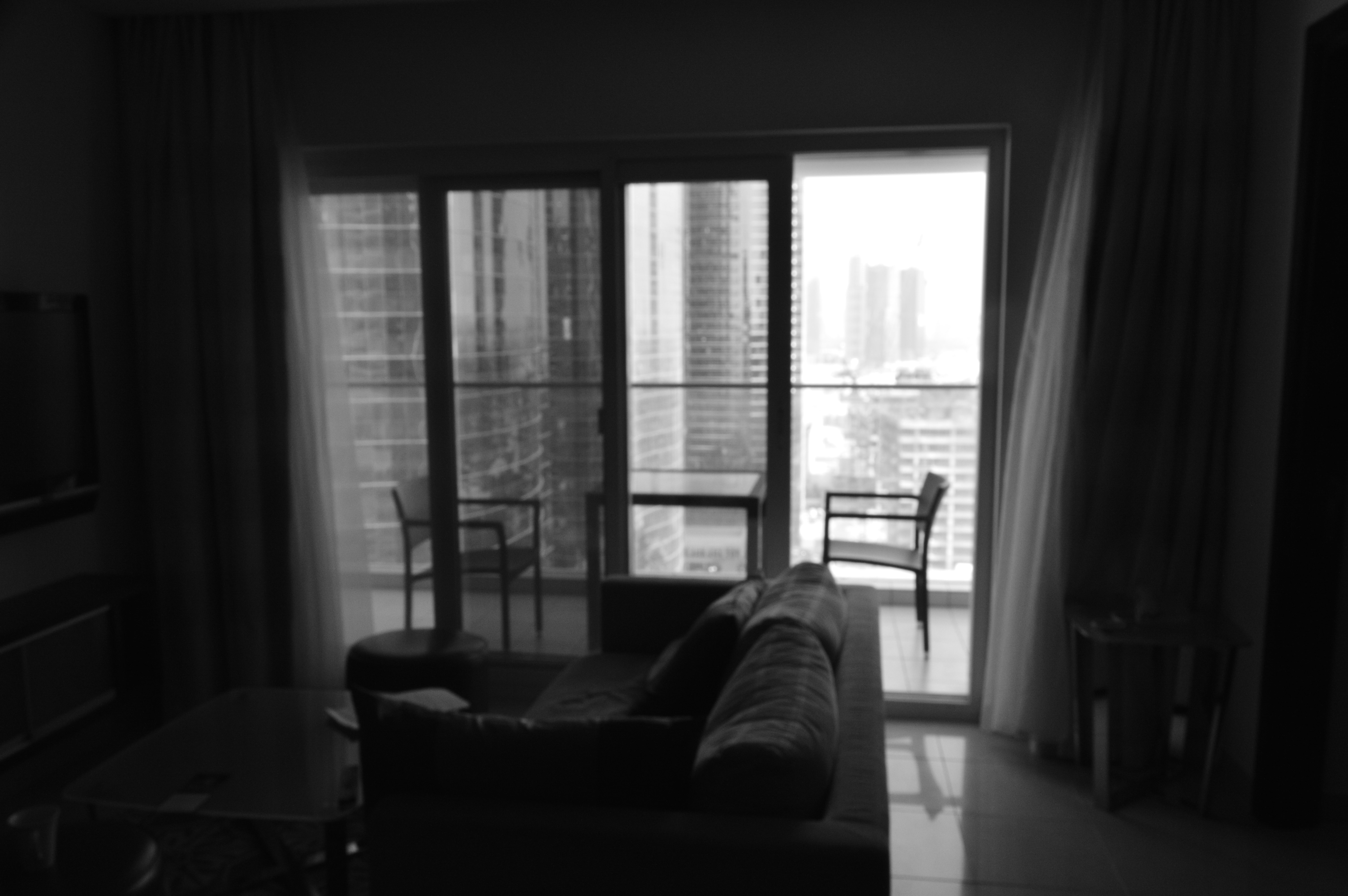 The image depicts a cozy indoor setting with a person lying on a couch, facing a large window or glass door that opens to a balcony. The room is dimly lit, with natural light streaming in from the window. The balcony has a table and chairs, suggesting a space for relaxation or enjoying the view. The overall atmosphere appears calm and serene.
