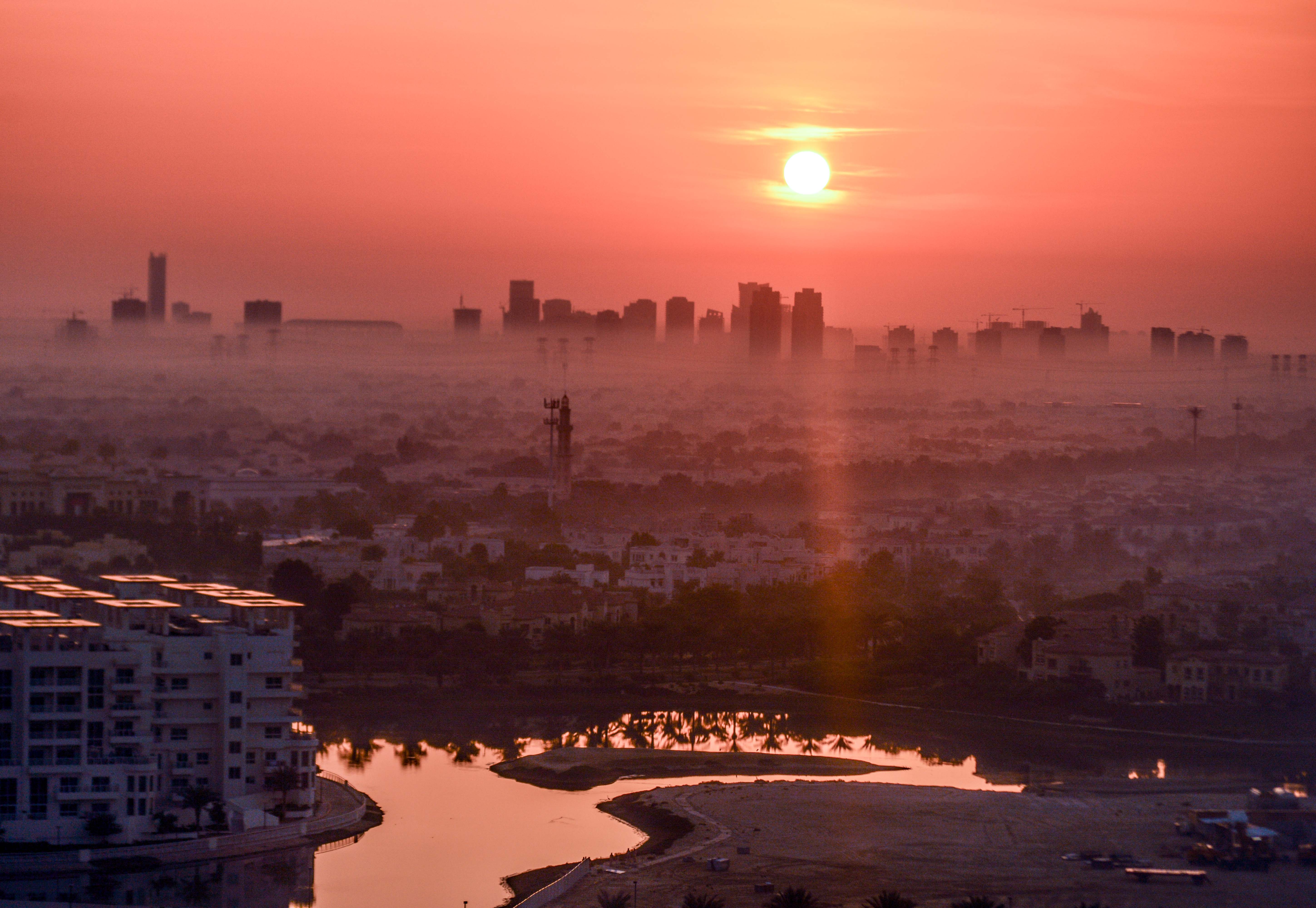 The image depicts a cityscape at sunrise or sunset, with the sun positioned low on the horizon, casting a warm, orange glow over the scene. The city is densely populated with buildings of varying heights, and the skyline is somewhat hazy. In the foreground, there is a body of water reflecting the sunlight, surrounded by a mix of residential and commercial structures. The overall atmosphere is serene and tranquil, typical of early morning or late evening.