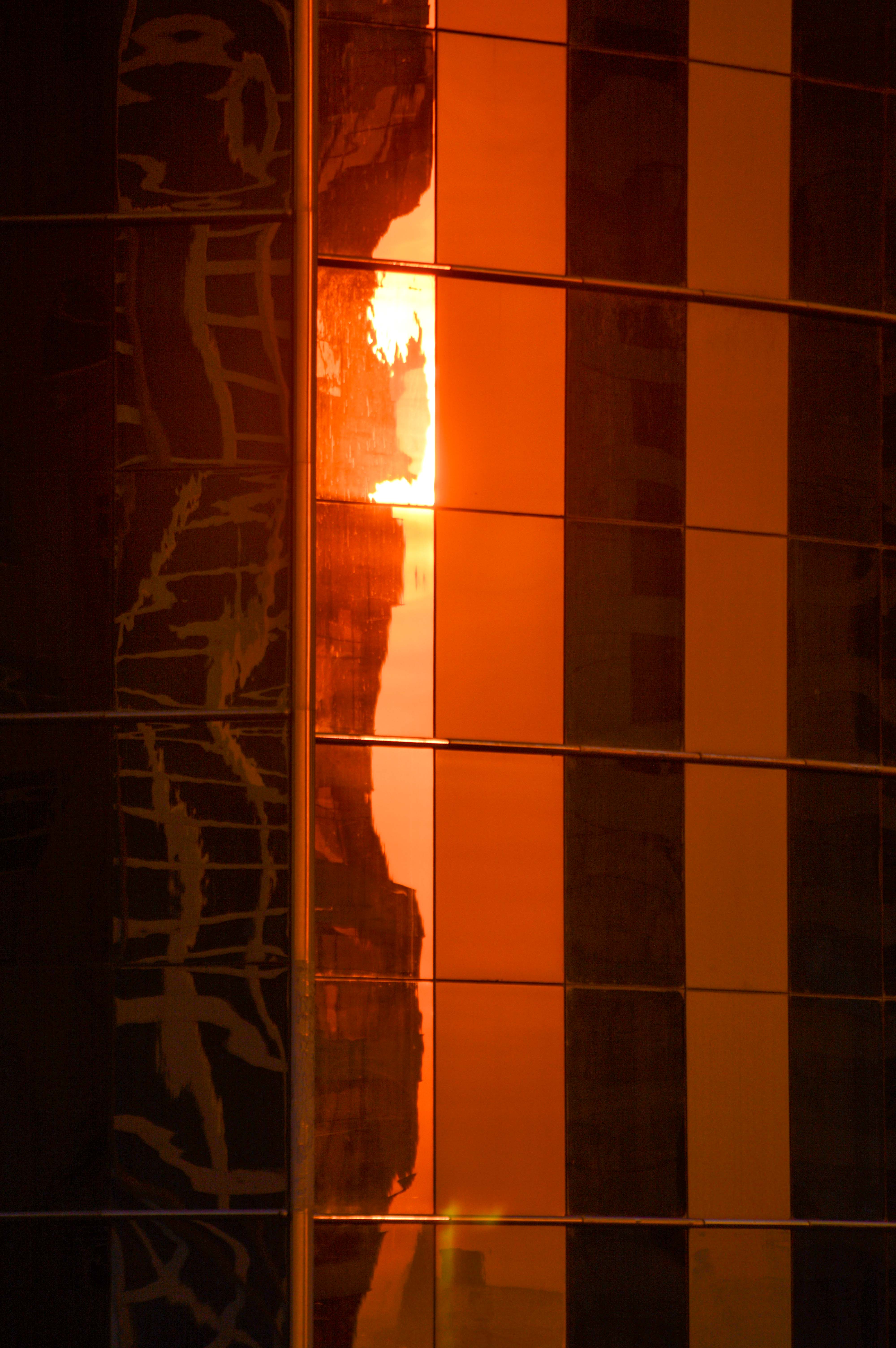 The image shows a reflection of a sunset or sunrise on a series of vertical, metallic, rectangular panels. The reflection creates a warm, glowing effect with a gradient of orange and yellow hues. The panels are arranged in a grid pattern, and the reflection captures the light in a way that highlights the texture and lines of the panels. On the left side, there is a dark, abstract pattern that contrasts with the bright reflection on the right side.
