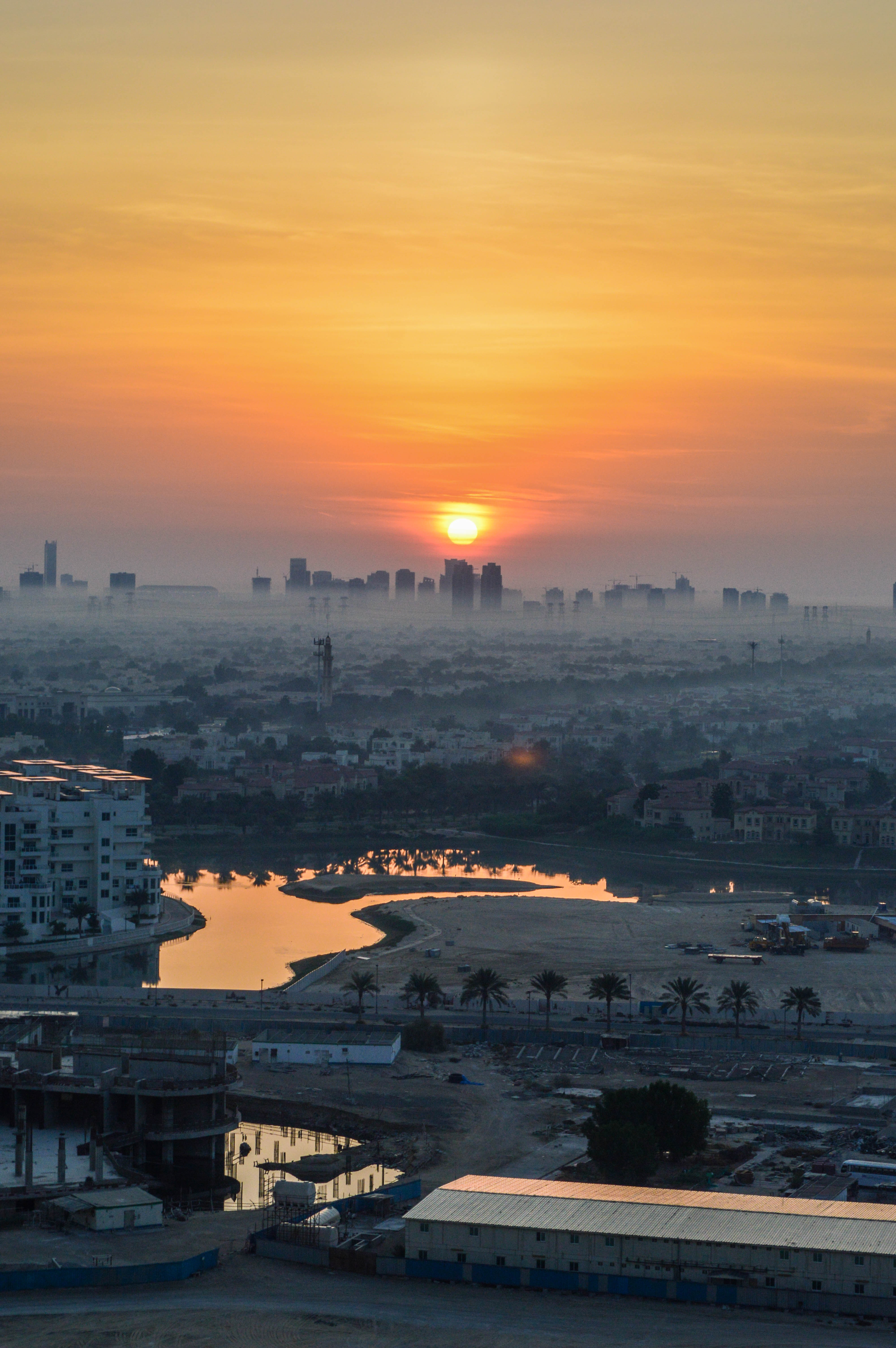 The image depicts a cityscape at sunset, with the sun setting behind a skyline of buildings. The foreground features a mix of residential and industrial structures, including a partially constructed building, a water body reflecting the warm hues of the sunset, and several palm trees. The overall scene is bathed in the golden light of the setting sun, creating a serene and picturesque atmosphere.