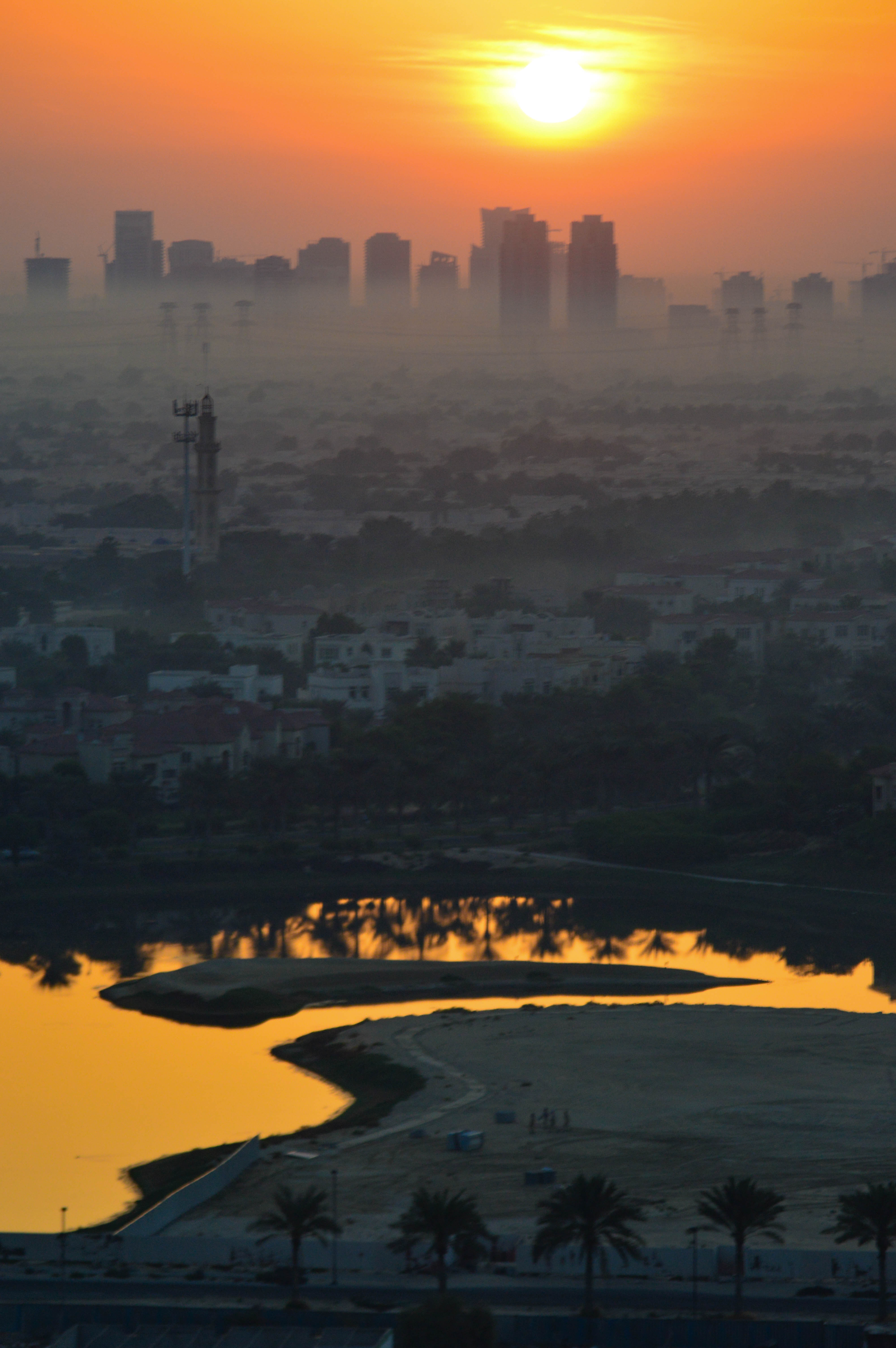 The image depicts a cityscape at sunset, with the sun low on the horizon casting a warm, orange glow over the scene. In the foreground, there is a winding body of water reflecting the sunlight, surrounded by sandy areas and a few palm trees. Beyond the water, a residential area with numerous houses and buildings can be seen. In the background, the skyline of the city is visible, featuring several tall buildings and skyscrapers. The overall atmosphere is serene and tranquil, capturing the beauty of the city at sunset.