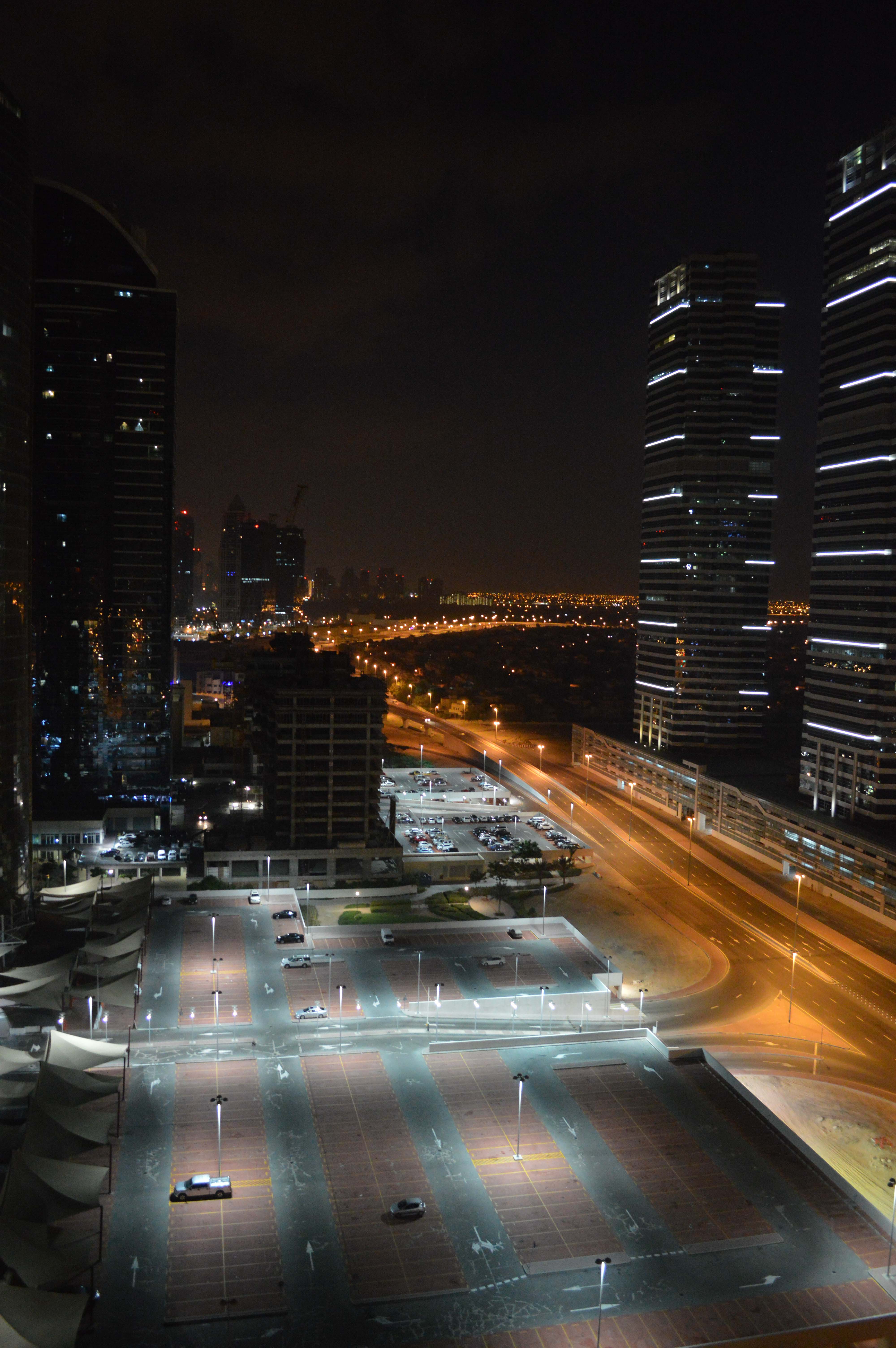 The image depicts a nighttime cityscape featuring tall, illuminated skyscrapers and a well-lit parking lot with sparse vehicular traffic. The scene includes a mix of modern high-rise buildings and a few shorter structures, with a highway in the background. The overall atmosphere is calm and orderly, with a clear view of the city's infrastructure and layout.
