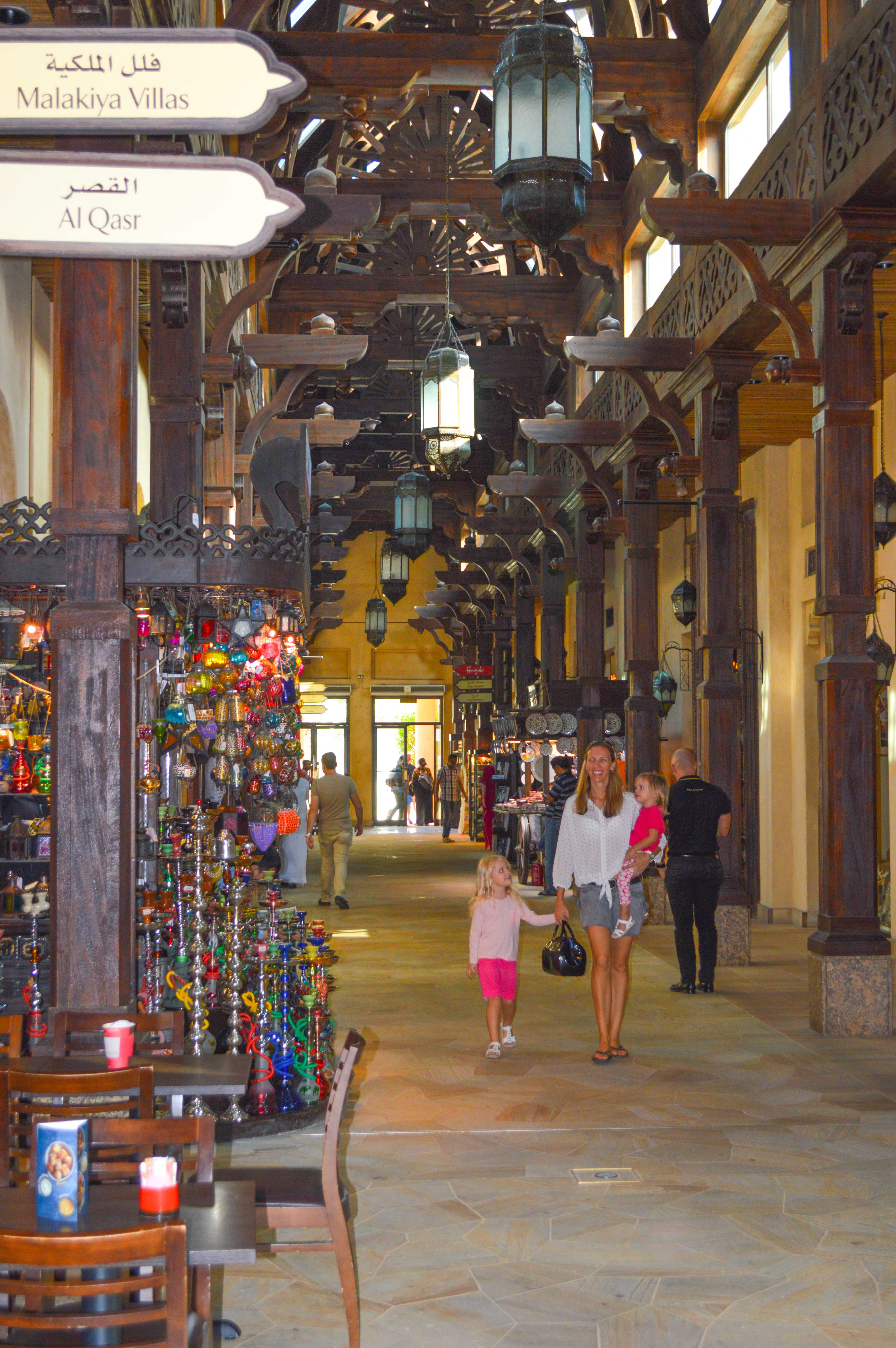 The image depicts a shopping area with a traditional architectural style, featuring wooden beams, lanterns, and signs in both Arabic and English. The signs indicate directions to 'Malakiya Villas' and 'Al Qasr'. The area is bustling with people, including a woman holding a child and another child walking in front of them. Shops on the left display colorful glassware and various items for sale. The overall atmosphere suggests a market or souk setting, likely in a Middle Eastern location.