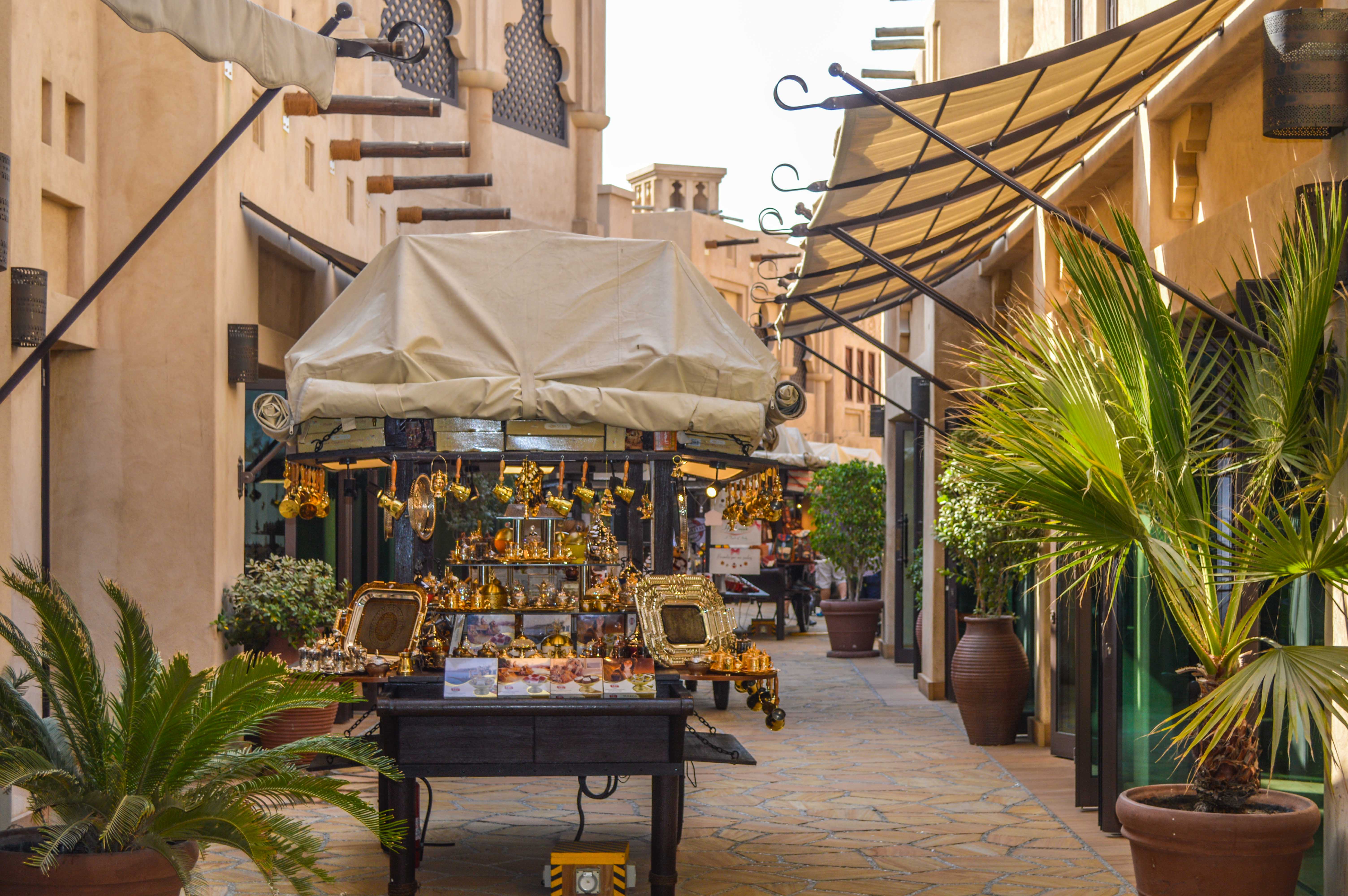 The image depicts a narrow, outdoor marketplace with a Middle Eastern or Mediterranean architectural style. The market features a variety of gold-colored items, including decorative frames and other ornaments, displayed on tables under a canopy. The area is adorned with potted plants and has a tiled pathway, creating a charming and inviting atmosphere.