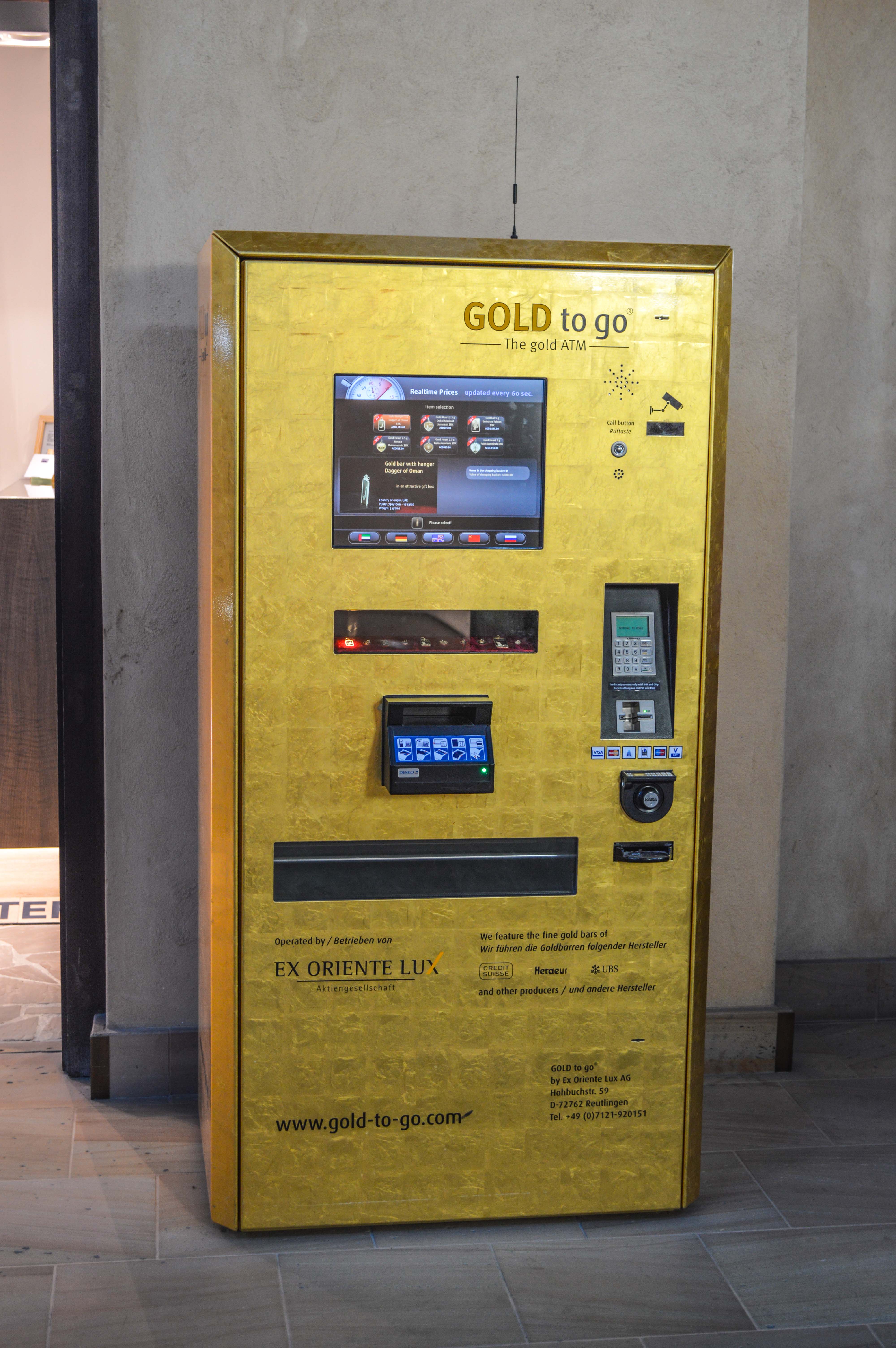 The image shows a gold ATM machine labeled 'GOLD to go' operated by Ex Oriente Lux AG. It allows users to purchase gold bars with real-time pricing updated every 60 seconds. The machine features gold bars from various producers like Credit Suisse, Heraeus, and LIBS. Instructions and contact information are provided on the machine, and it is accessible via the website www.gold-to-go.com.